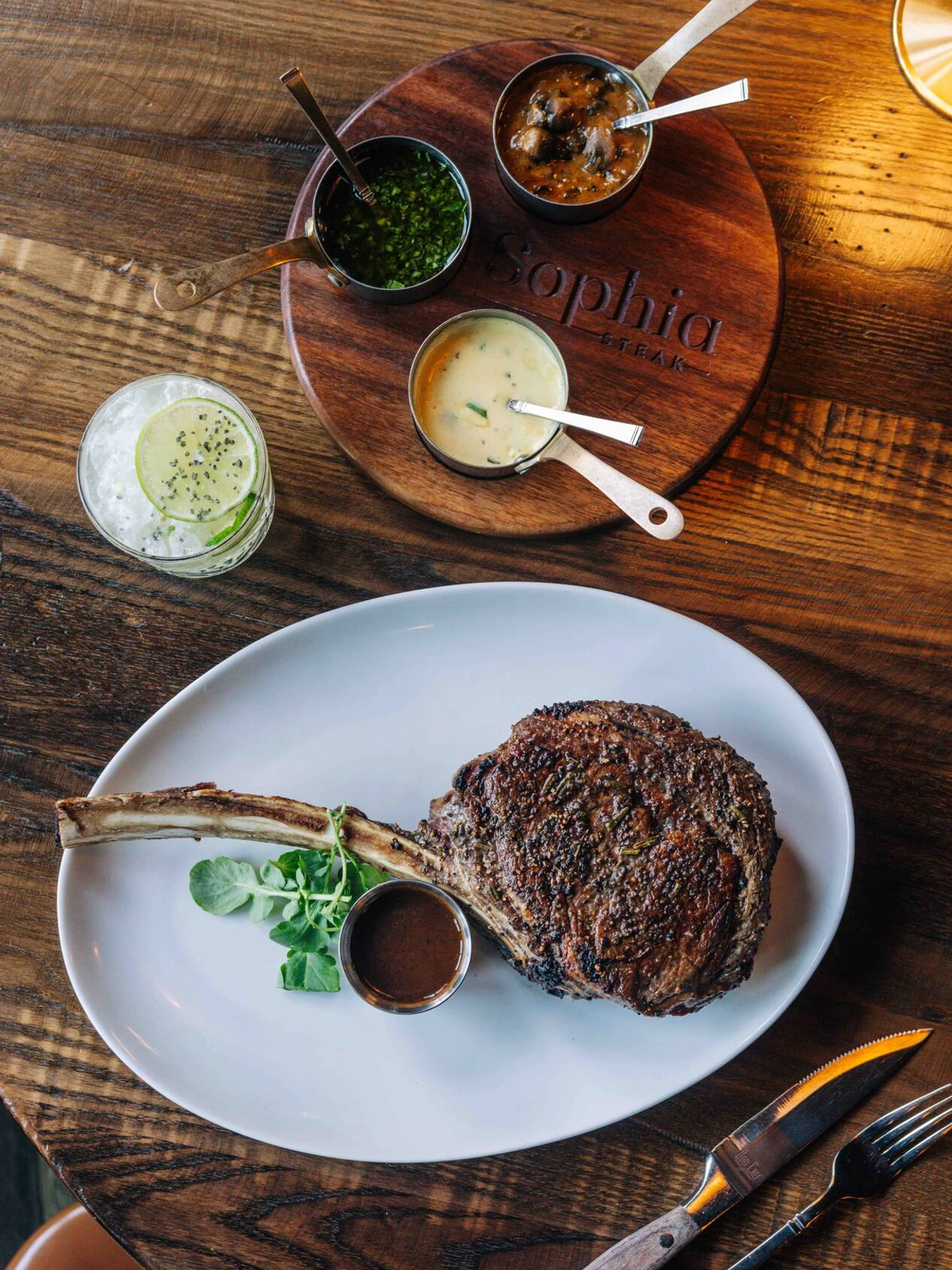 Grilled steak with sauce on white plate, three sauces in small bowls on wooden board, and a drink on table.