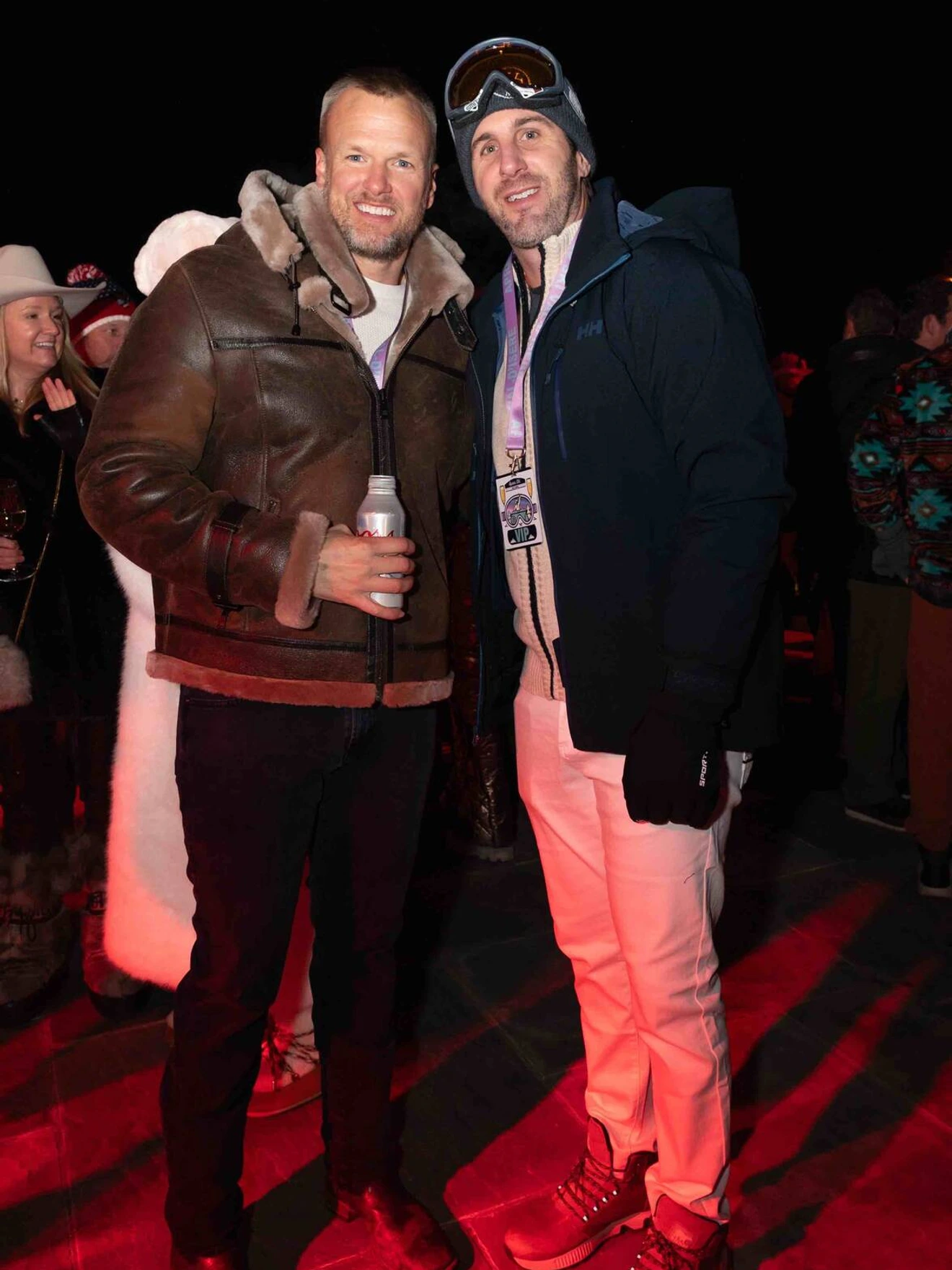 Two men in winter clothing posing at an outdoor nighttime event with other people in the background.