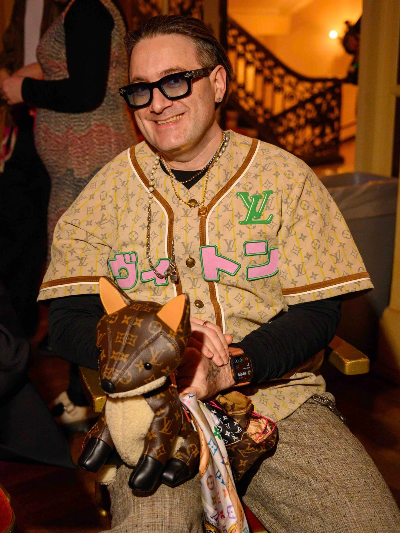 Man wearing patterned Louis Vuitton outfit and dark glasses, holding a designer dog plush toy in indoor setting.