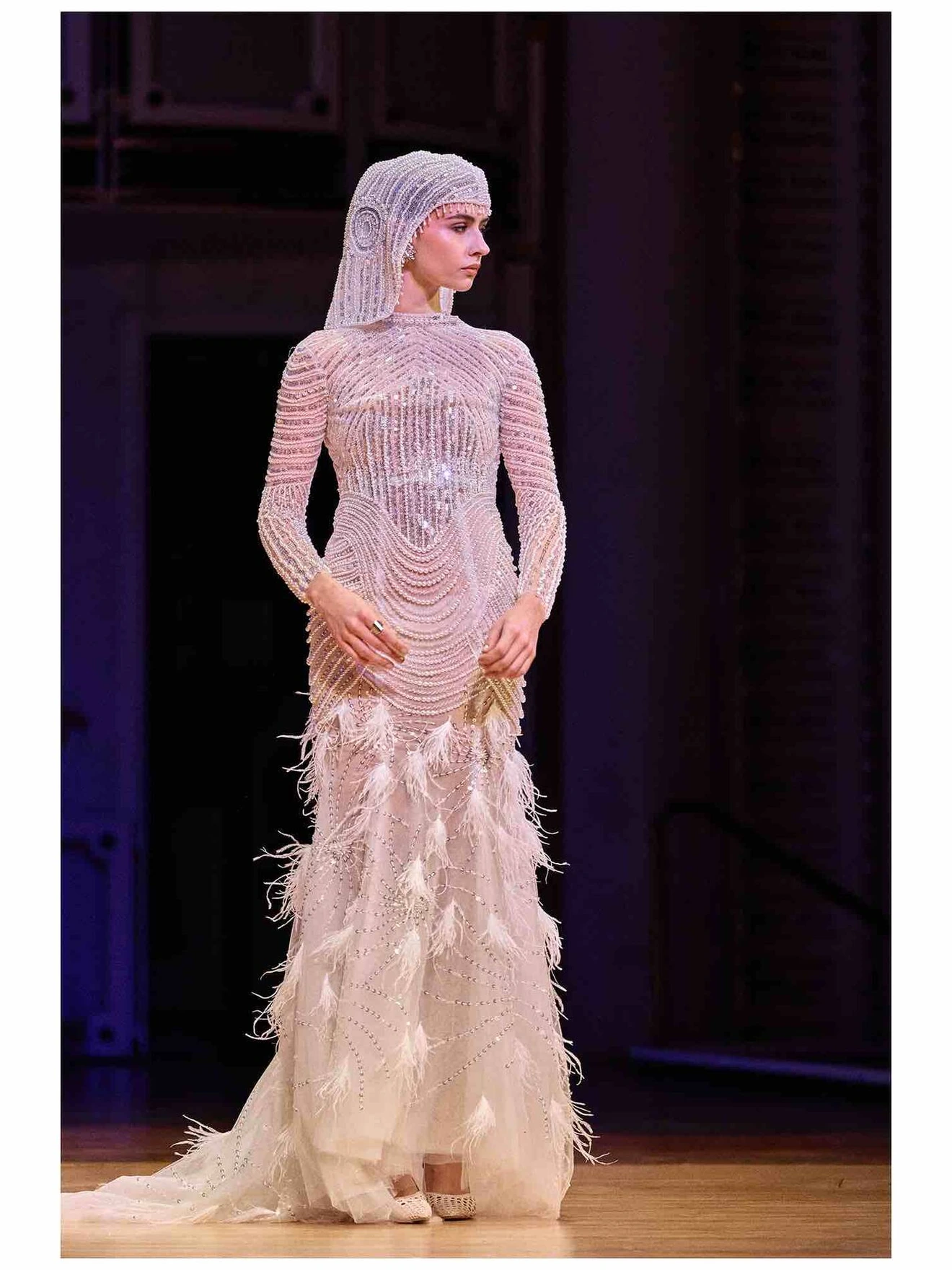 Model wearing an intricate, beaded white gown with feather details on a runway.