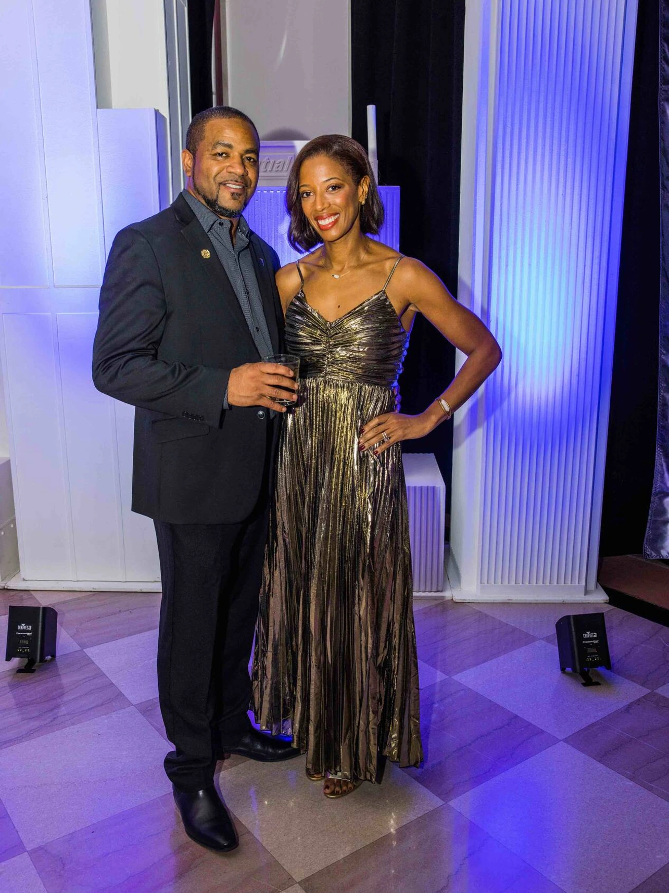 Man in dark suit and woman in metallic gown posing indoors with blue and purple lighting.