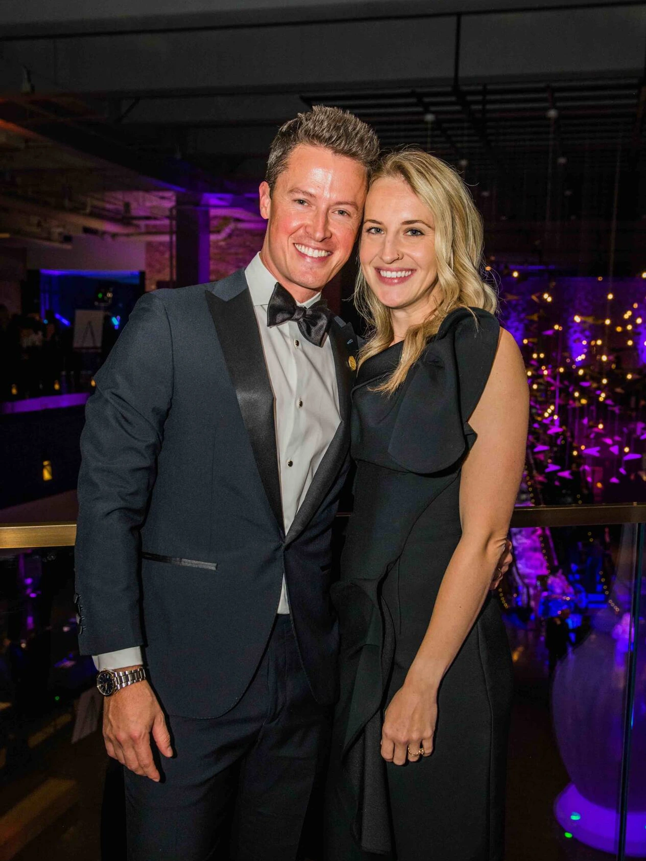 Man in tuxedo and woman in black evening dress posing together at an indoor event.