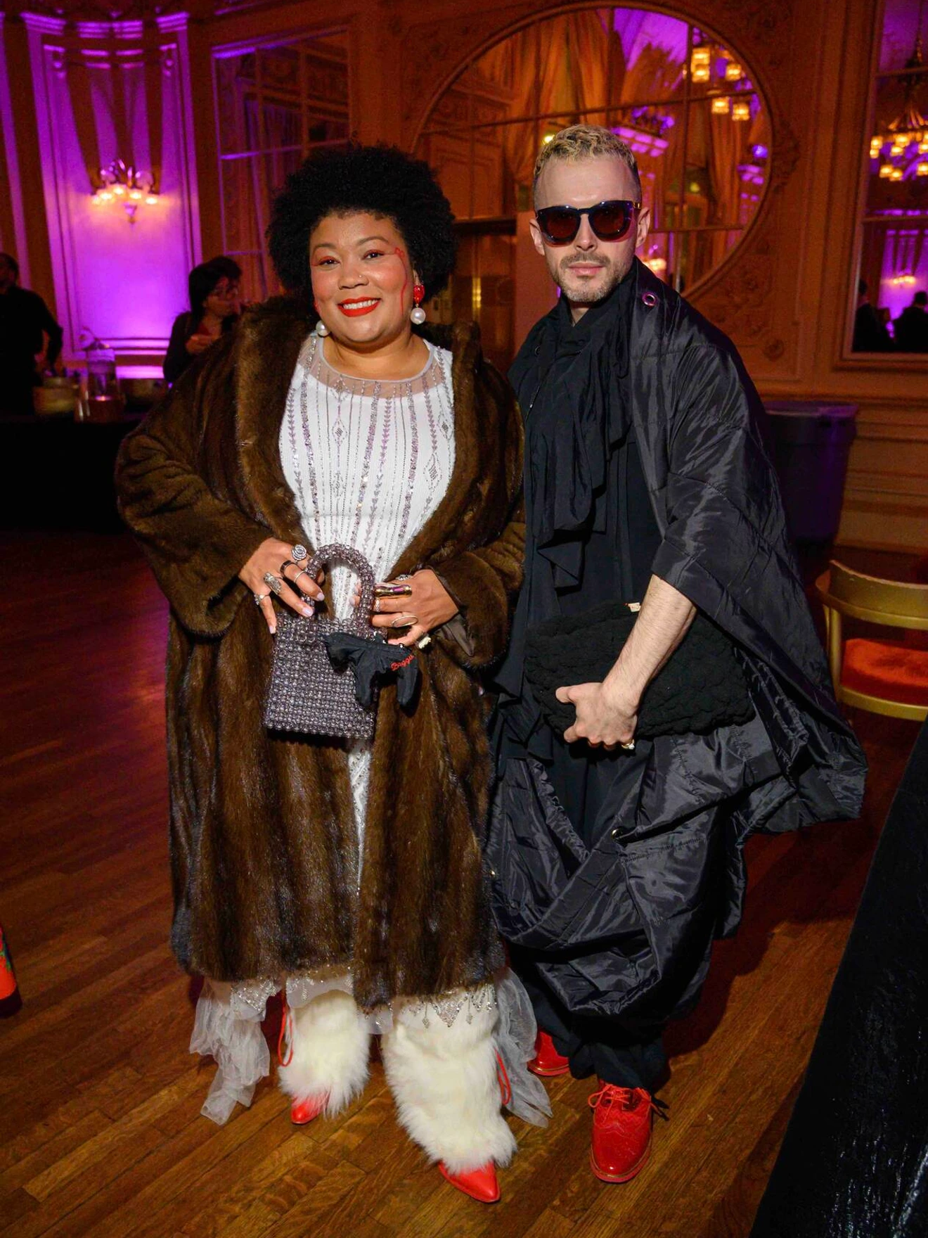 Two stylishly dressed people posing indoors at an event with purple lighting and ornate wood paneling.