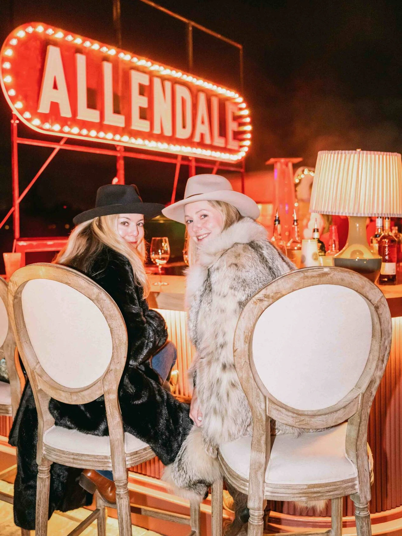 Two women in fur coats and hats sitting at a bar with a lit ALLENDALE sign in the background.