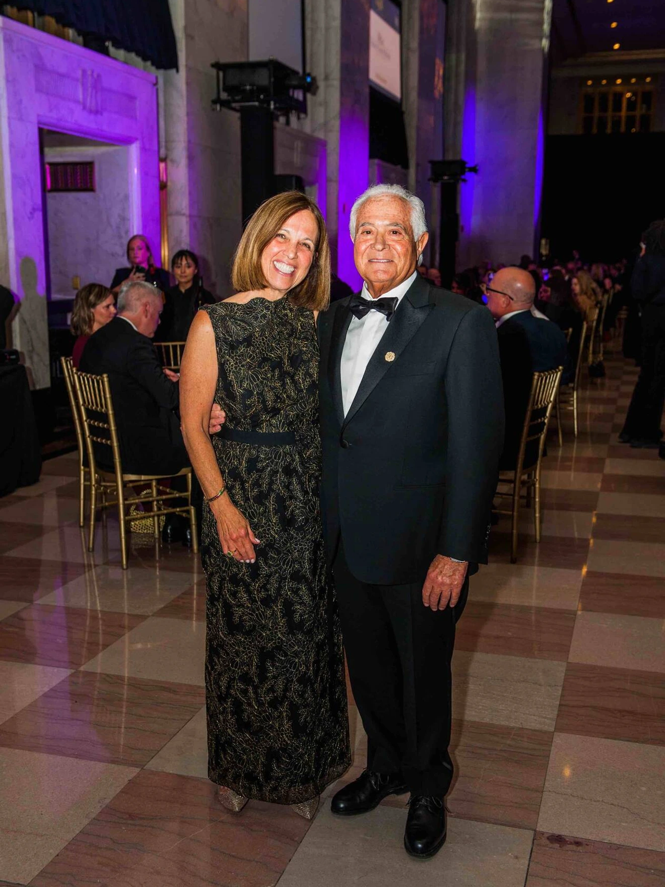 Man in tuxedo and woman in black dress posing at formal event with seated guests in background.