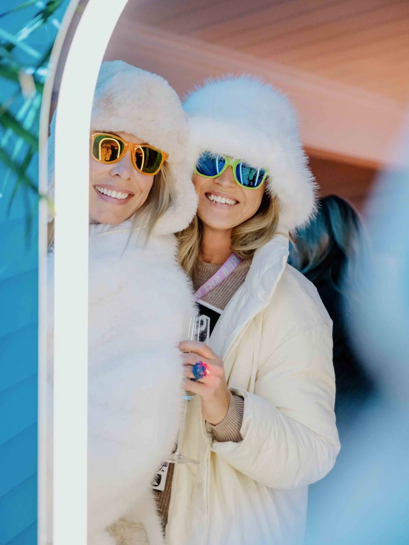 Two smiling people in white fur coats and sunglasses, posing indoors with a glass in hand.