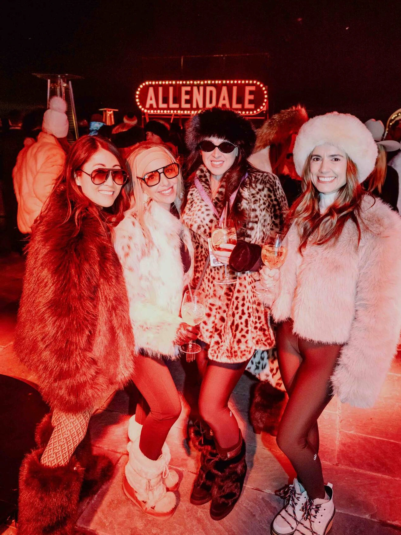 Four women in furry winter outfits and sunglasses posing together at a nighttime event with Allendale sign.