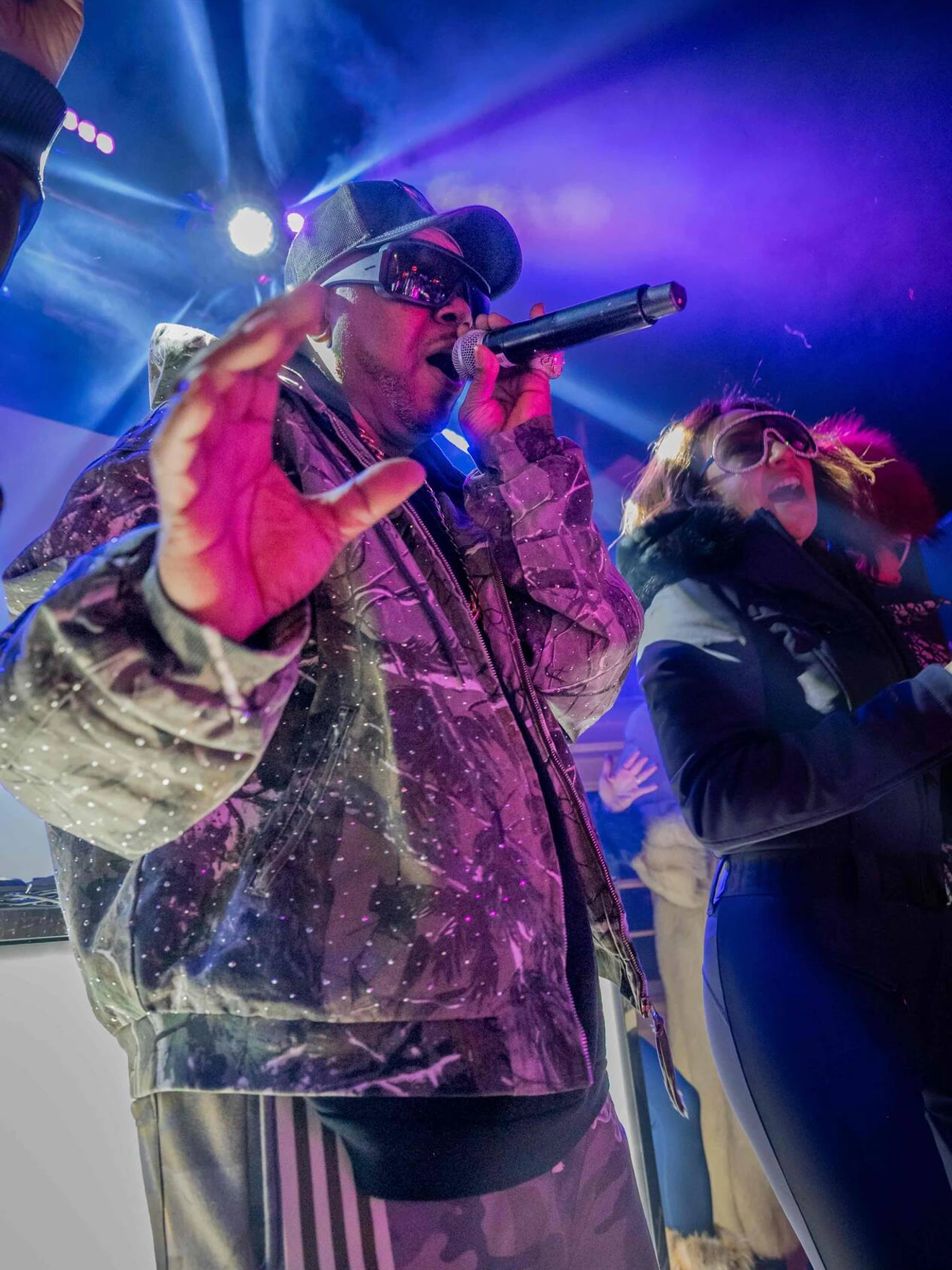 Man in camo jacket and cap rapping on stage with purple and blue lights, woman beside him dancing.