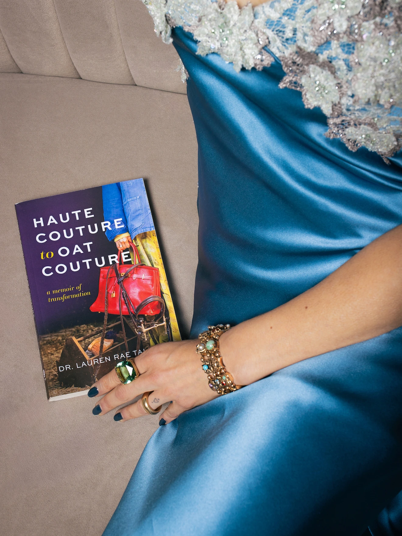 Hand wearing rings and bracelets rests on blue satin dress next to book titled Haute Couture to Art Couture by Dr. Lauren Raetz.