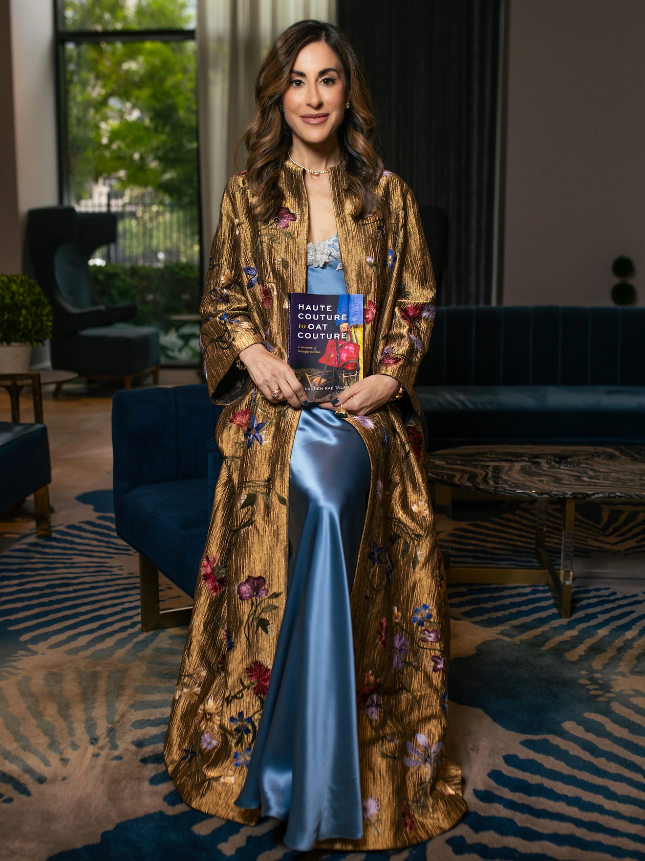 Woman in gold floral robe and blue satin dress sits on couch, holding a book titled Haute Couture + Goat Couture.