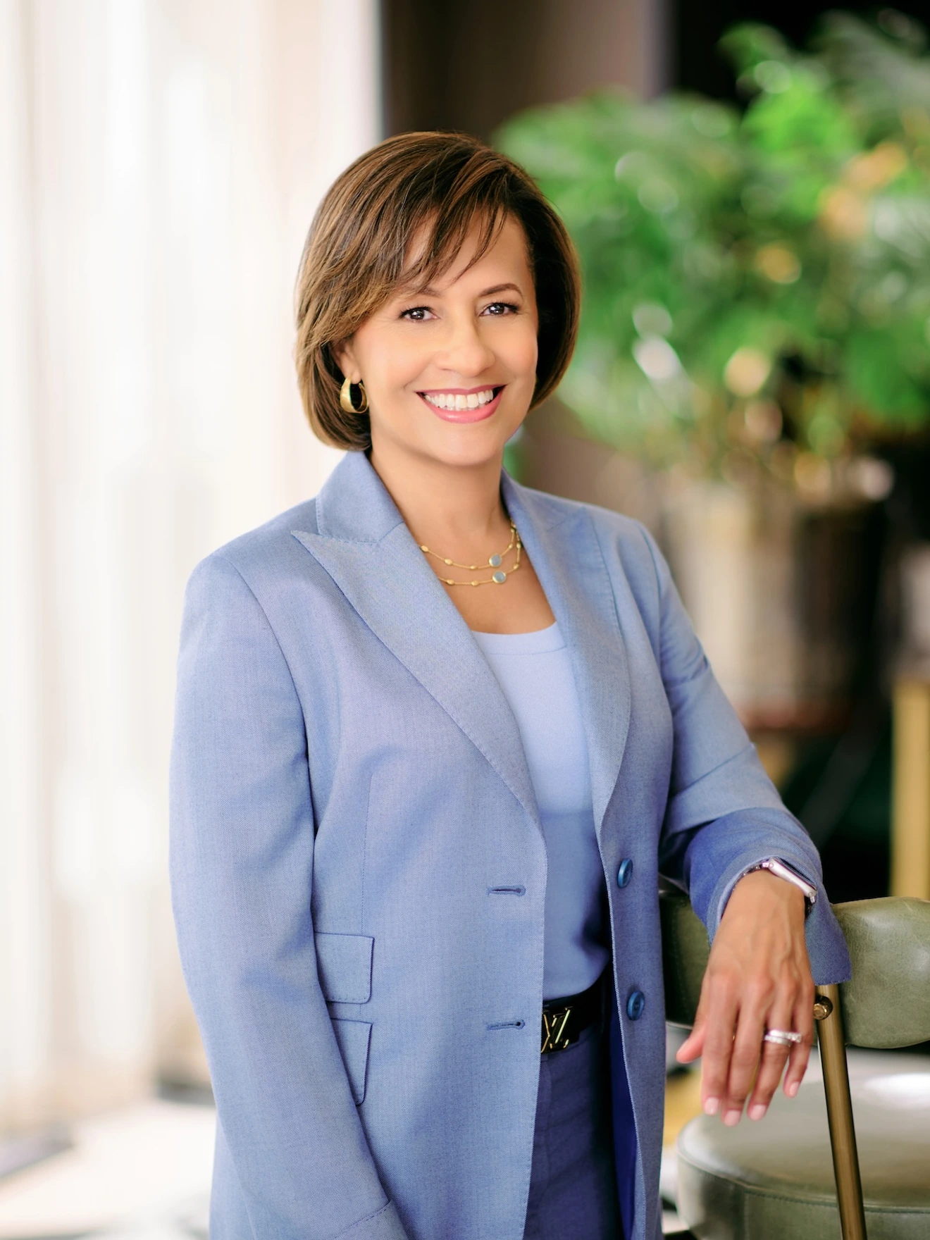 Smiling woman in a light blue suit leaning on a chair, with blurred plants in the background.