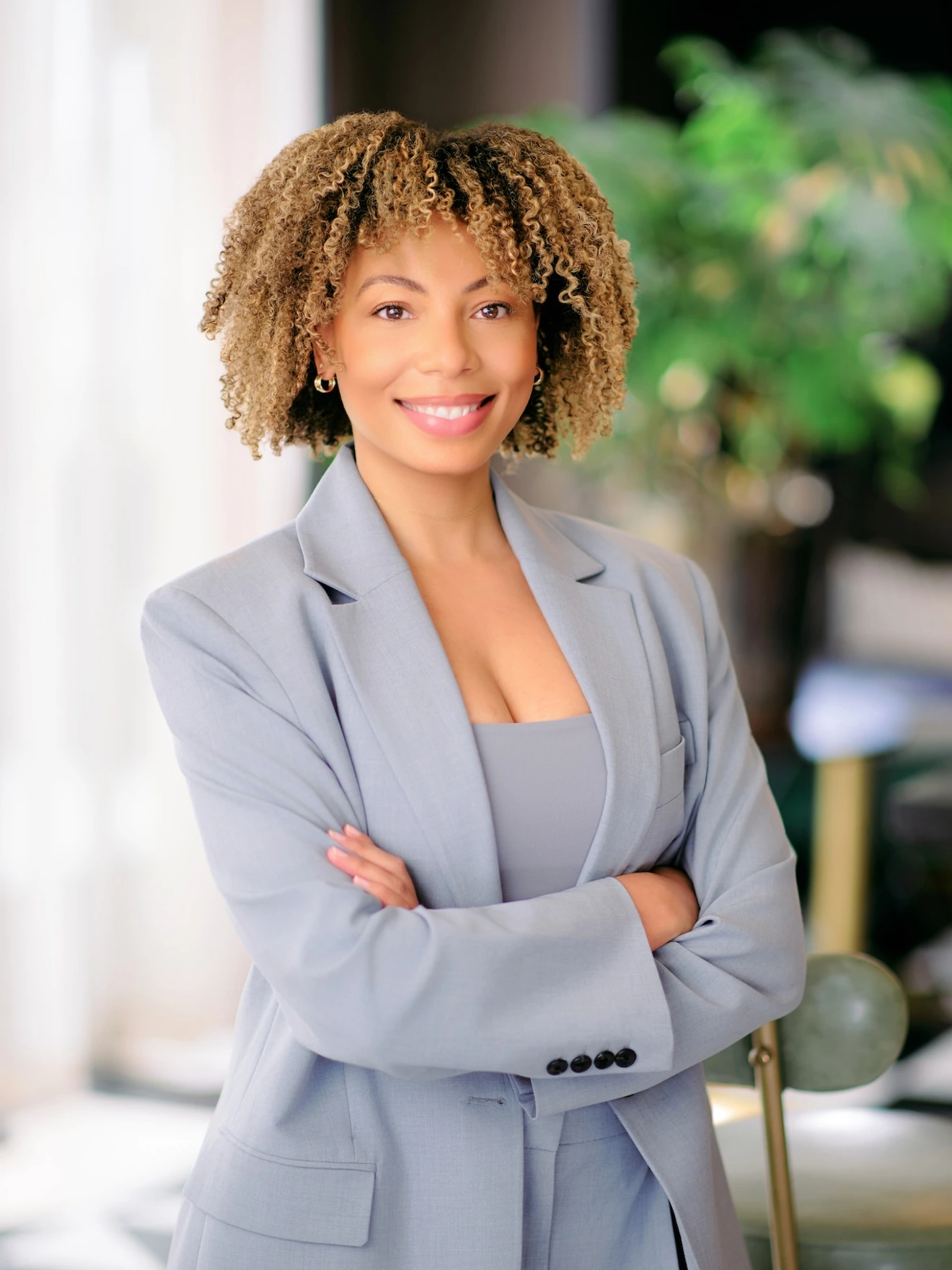Smiling woman with curly hair in light gray suit, arms crossed, standing in a softly lit indoor setting.
