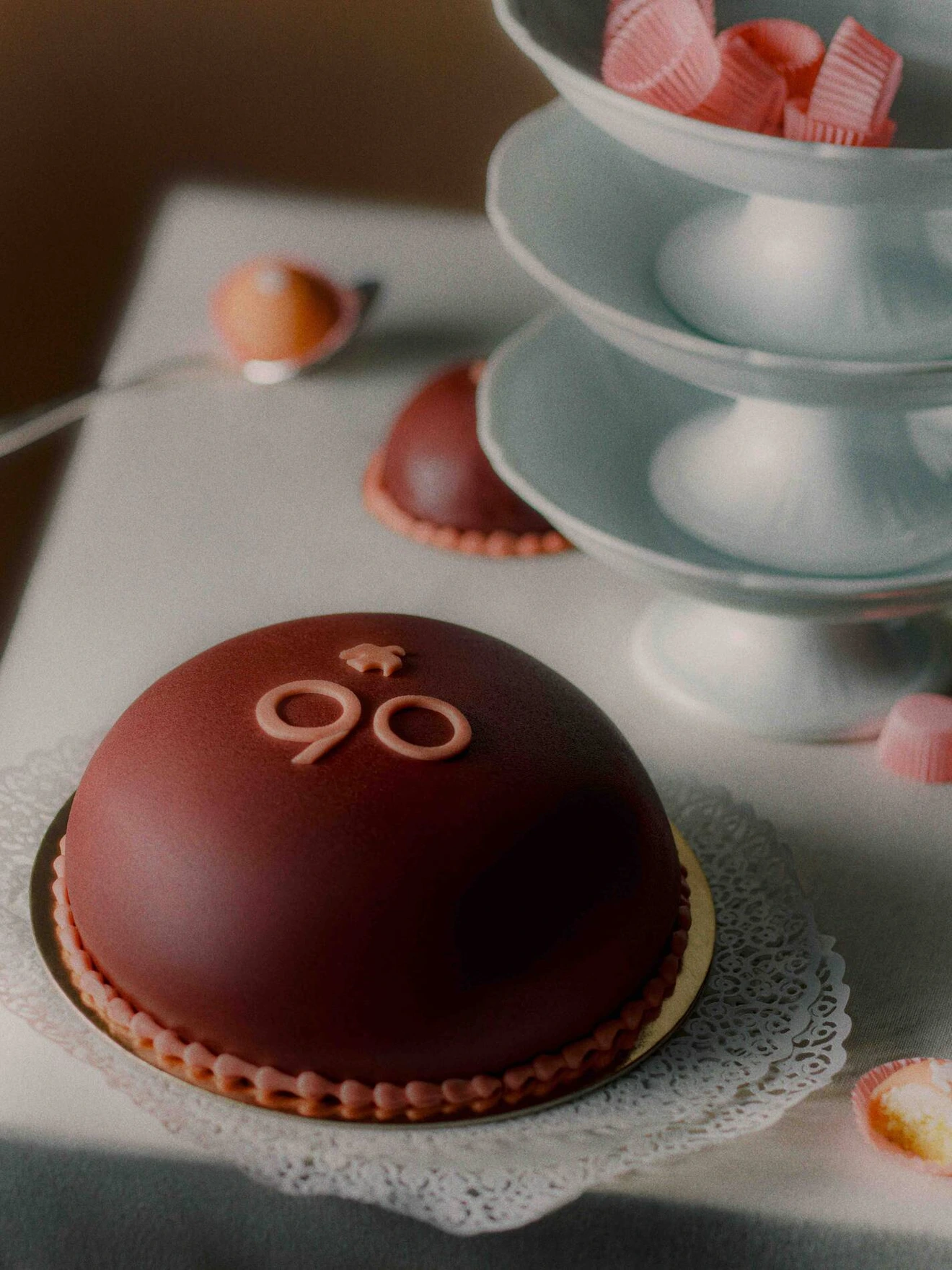 Round chocolate cake with 90 decoration on top beside a tiered tray with pink ribbon candies.