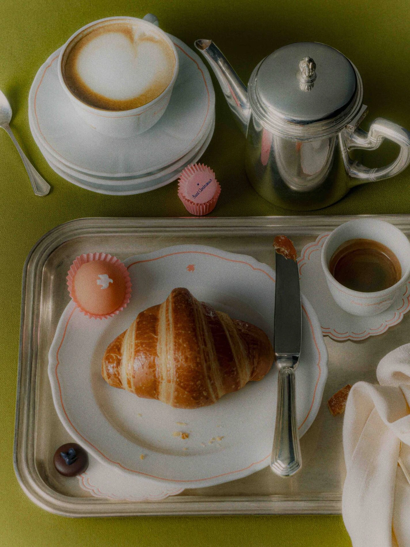 Silver tray with croissant, espresso, coffee pot, latte, and small pink confections on green table.