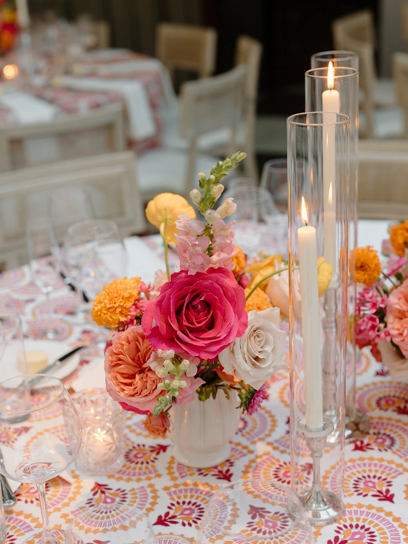 Table centerpiece with colorful flowers and tall glass candles on patterned tablecloth in dining setting.