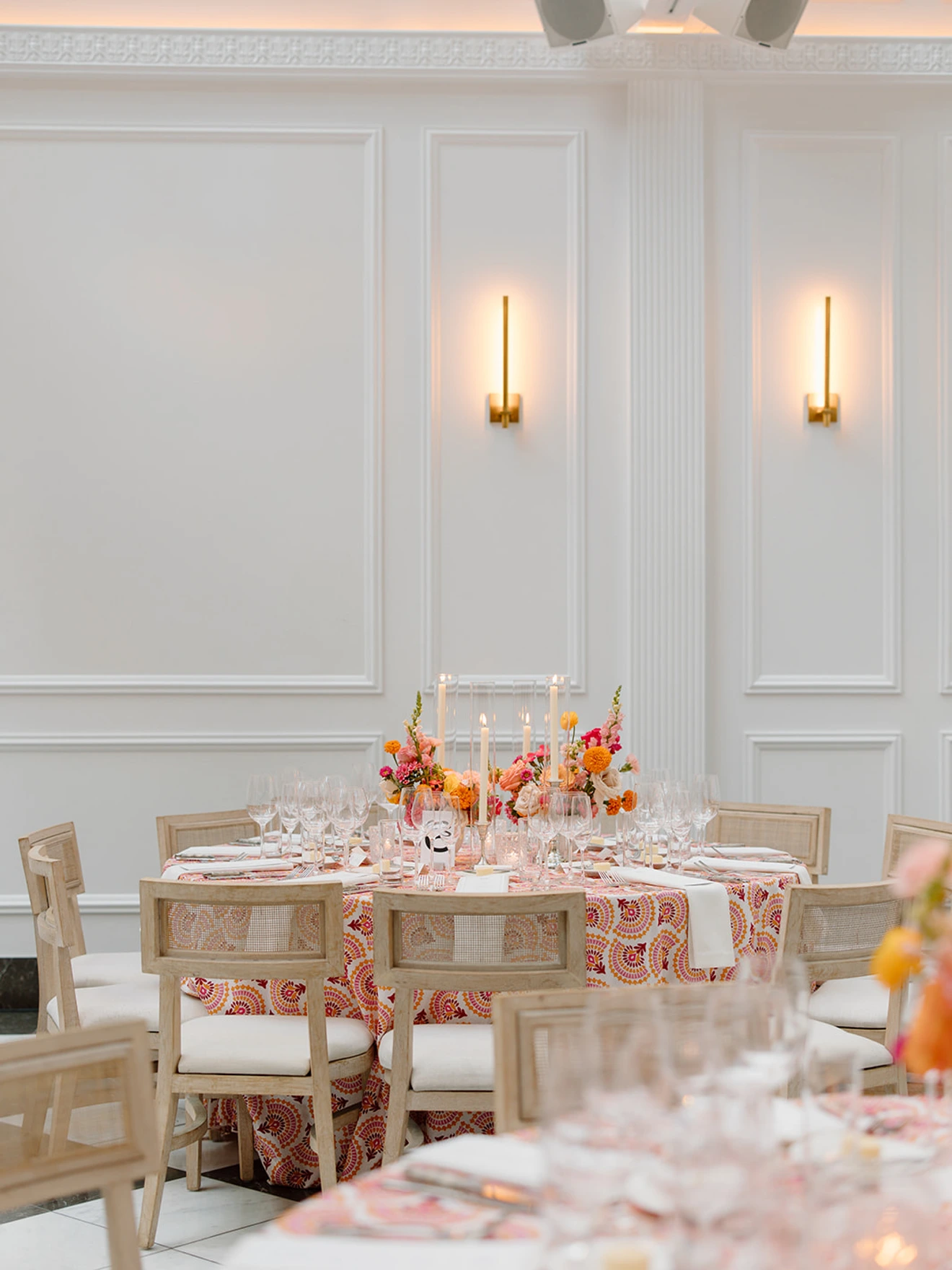 Elegant dining setup with floral centerpiece, candles, wooden chairs, and intricate tablecloth in a bright, white room.