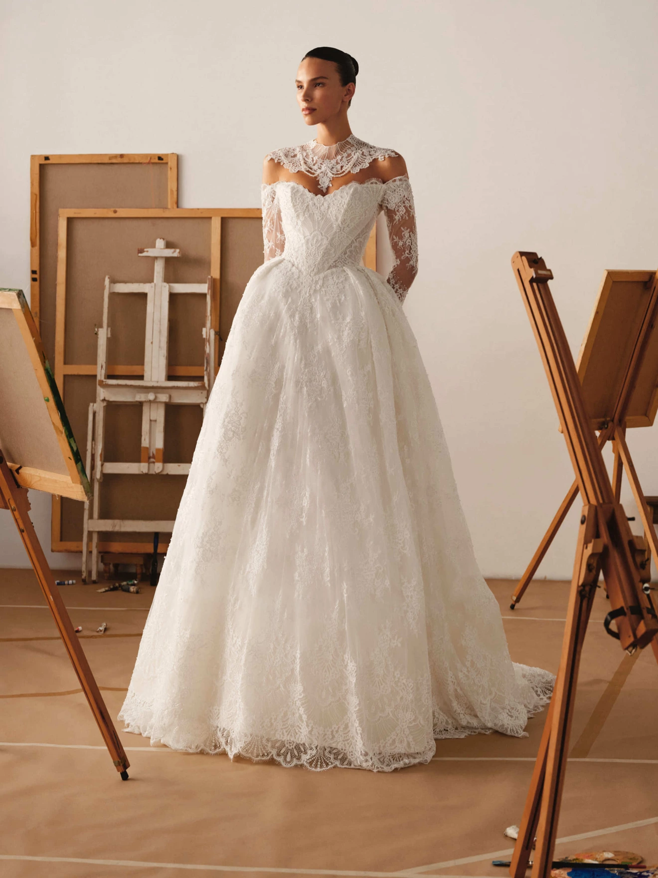 Woman in a white lace wedding dress standing in an art studio surrounded by easels and canvases.