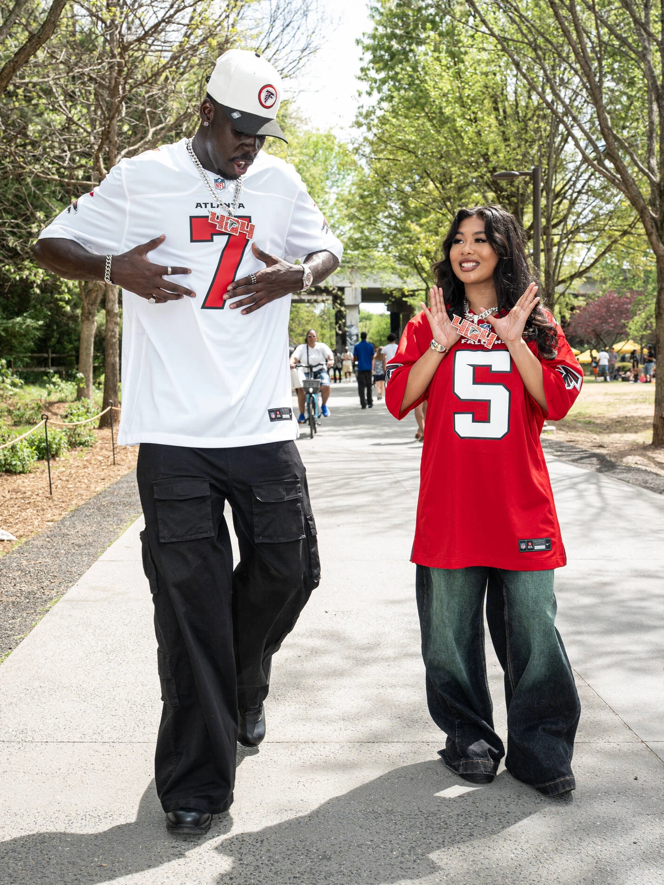 Two people wearing Atlanta Falcons jerseys, standing and gesturing on a sunny park pathway.