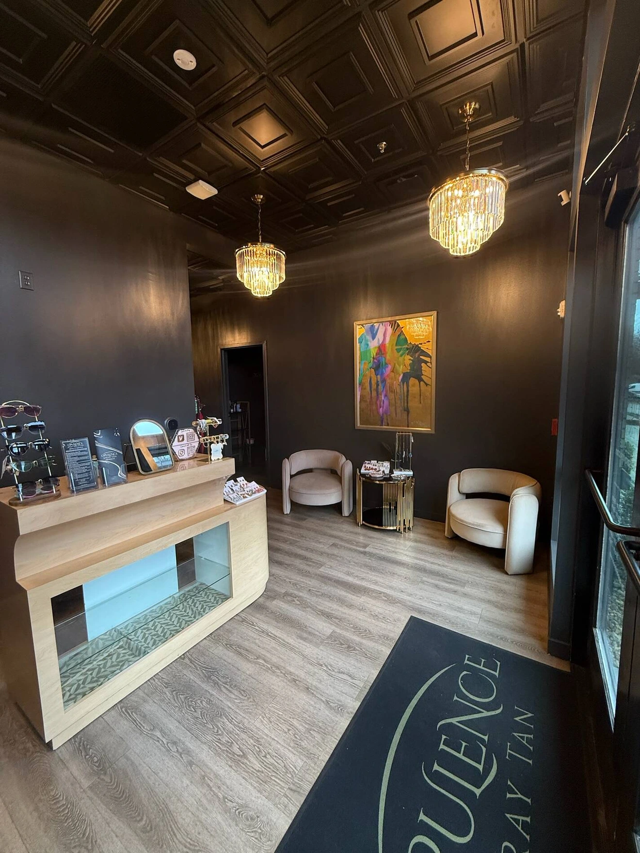 Modern, stylish tanning salon lobby with black walls, wooden floor, chandelier lights, counter, chairs, and colorful painting.