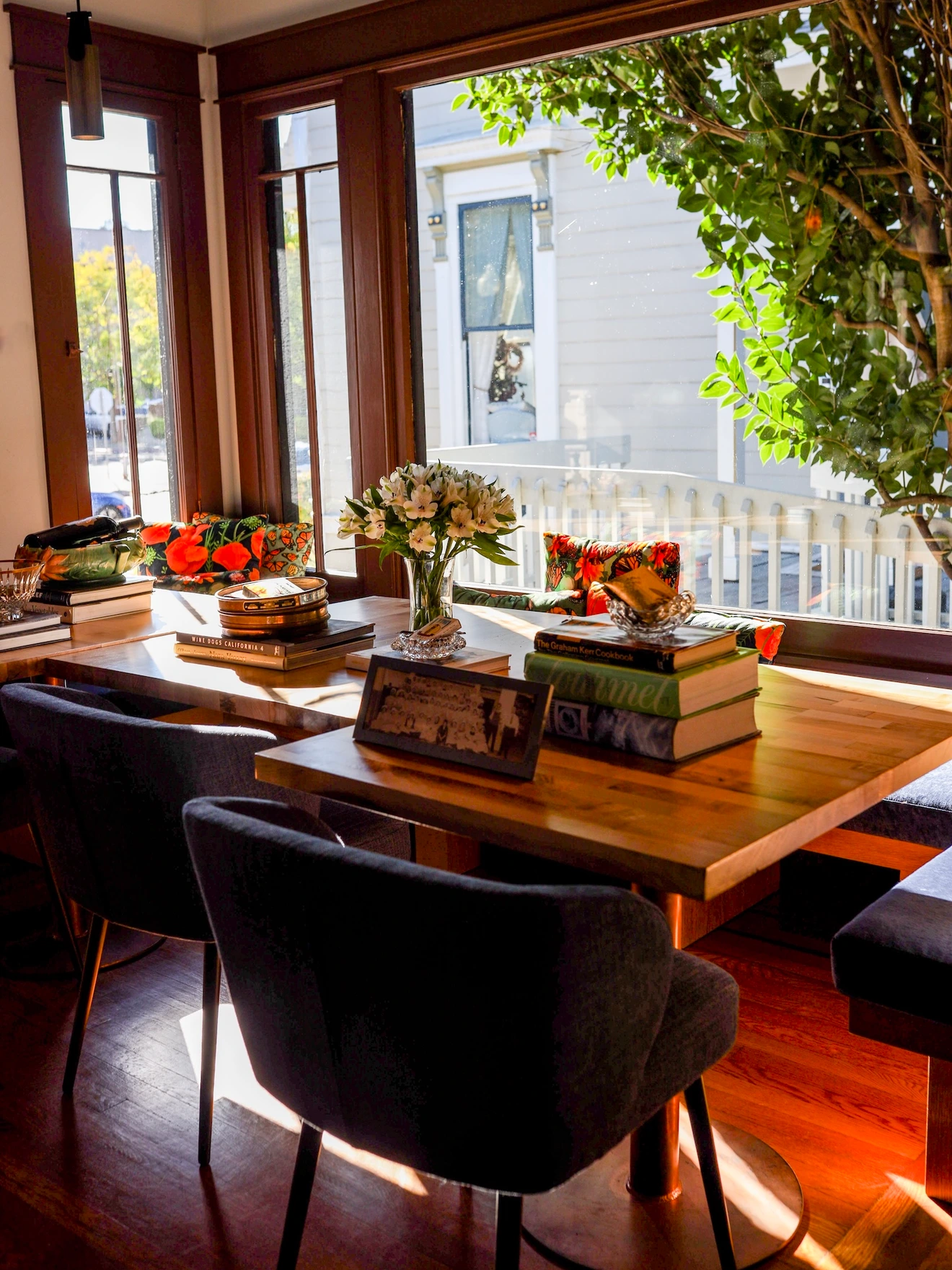 Sunlit wooden dining table with flowers, books, and chairs near large windows overlooking a porch and tree.