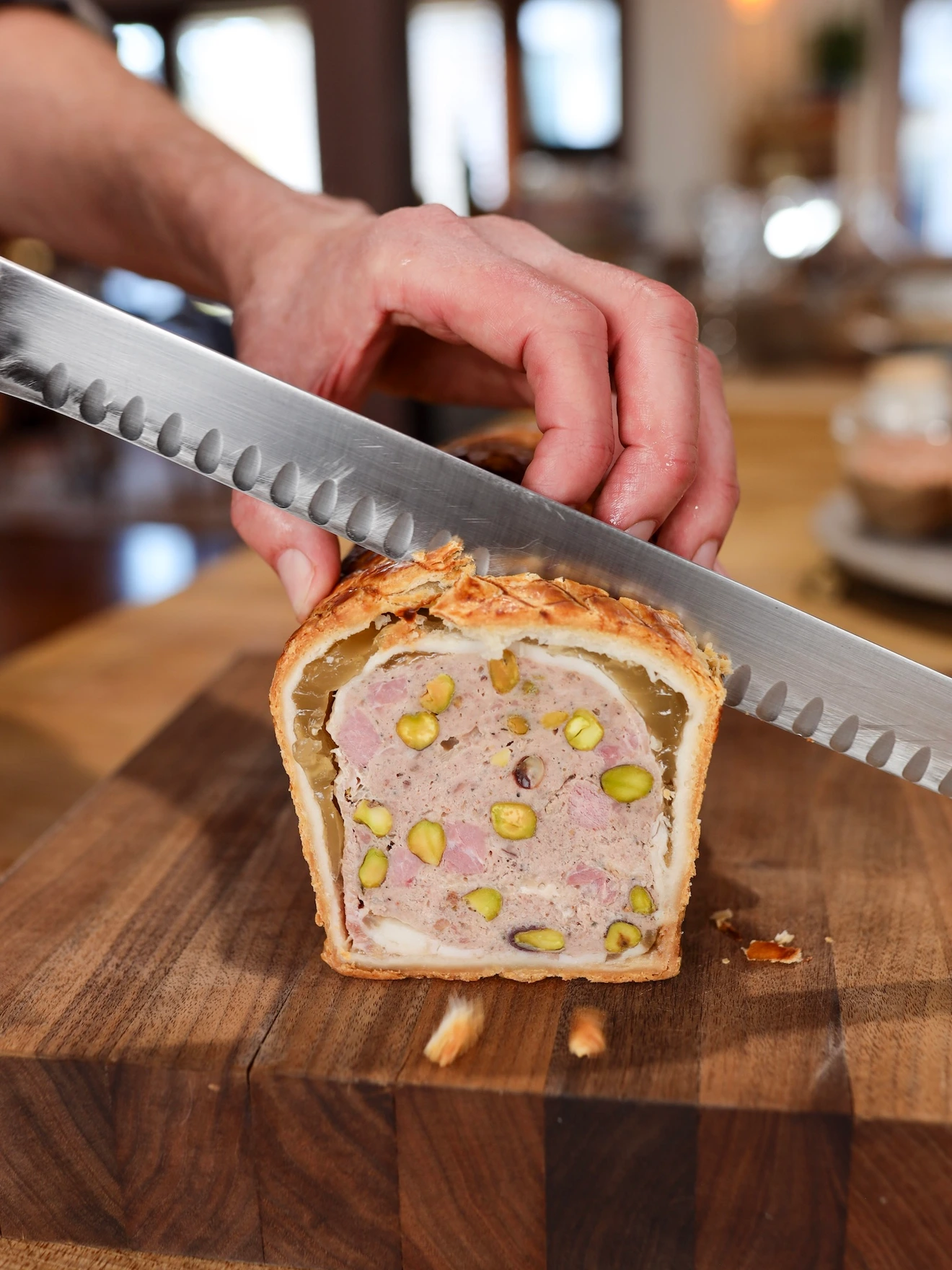 Hand cutting a slice of meat terrine with pistachios on a wooden board.