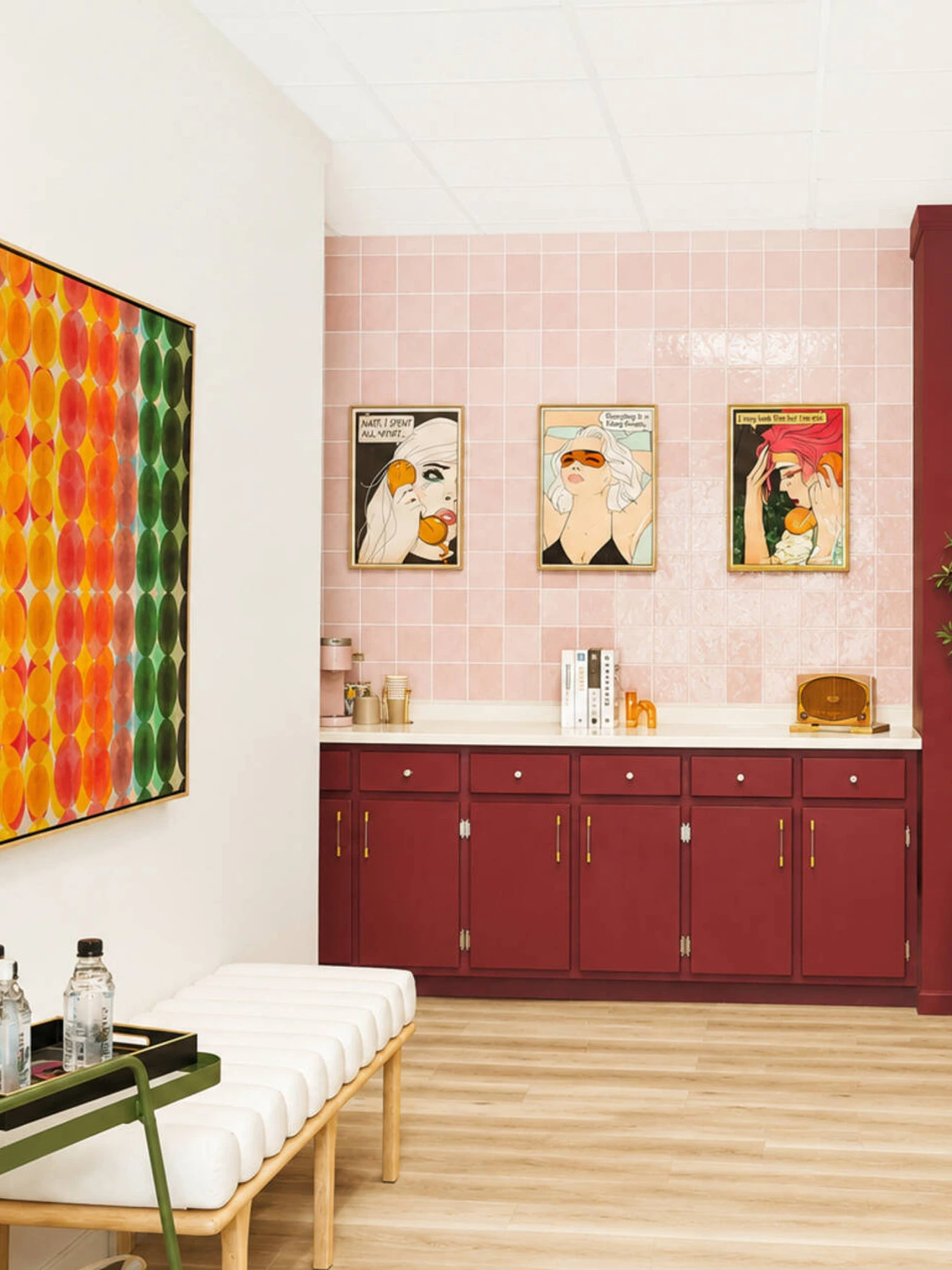 Modern waiting area with colorful artwork, red cabinets, white bench, water bottles, and a large potted plant.
