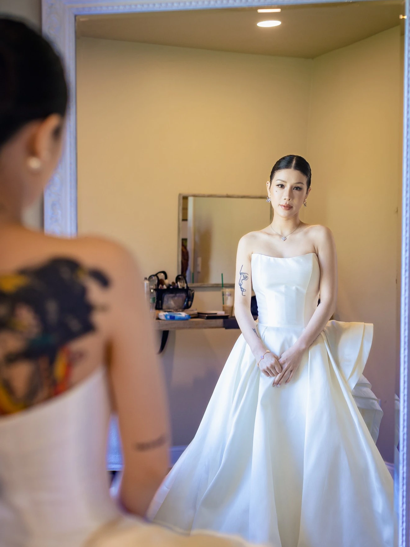 Woman in white strapless dress looking at herself in a large mirror in a softly lit room.