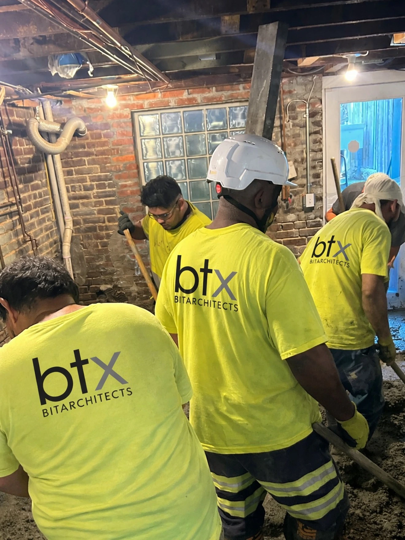 Four construction workers in yellow btx Bitarchitects shirts working in a basement with exposed brick walls.