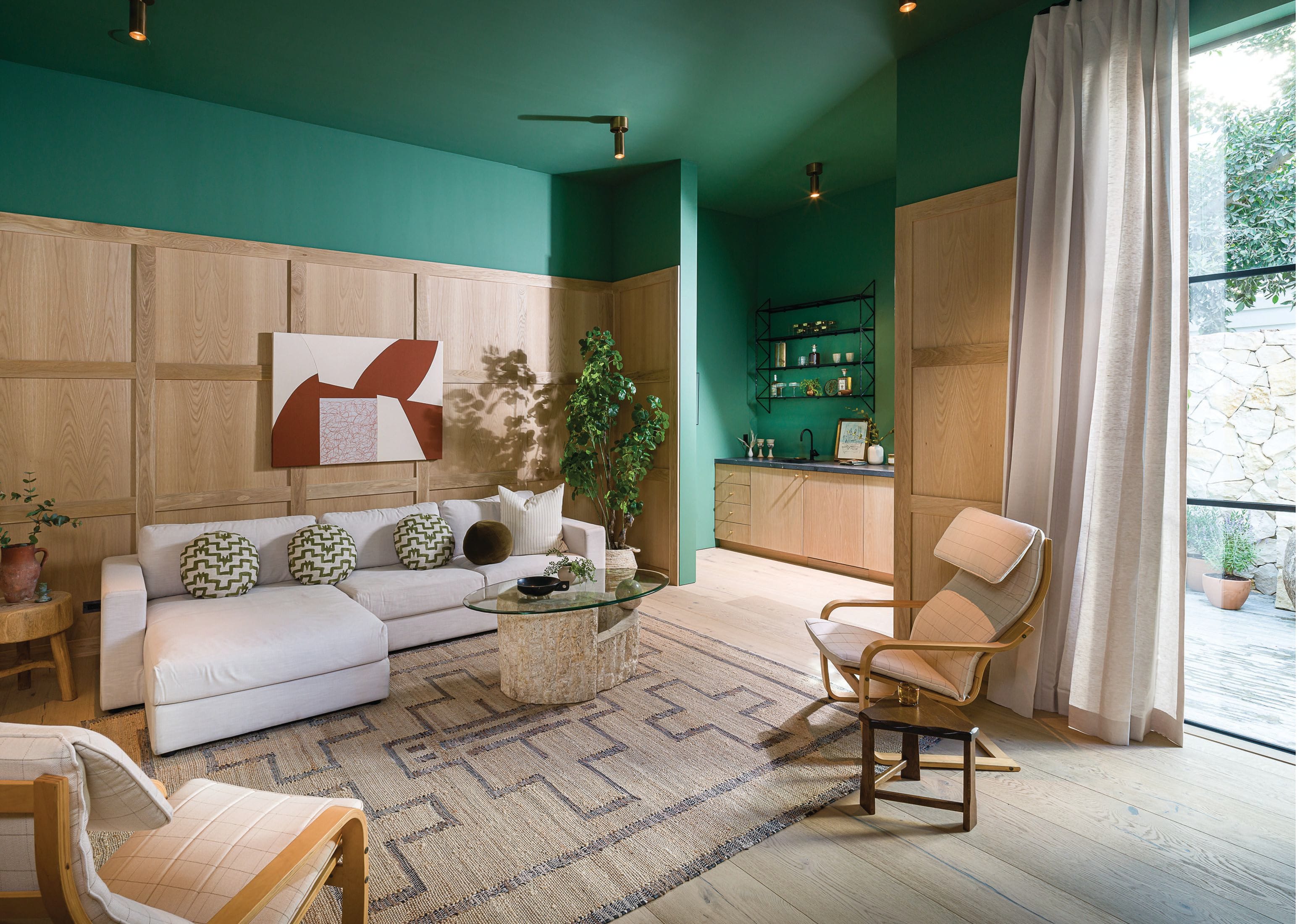 A bit moodier than the rest of the house, the den features walls covered in deepgreen paint by Farrow & Ball and a large white sectional adorned with round custom pillows made from Zak Fox fabric. The African-inspired Tuareg rug is from Burke Decor; the stone and glass coffee table is vintage; and the two Ikea Poäng chairs—reupholstered in a subtle windowpane print—add an unexpected sophisticated touch. PHOTOGRAPHED BY TODD GOODMAN