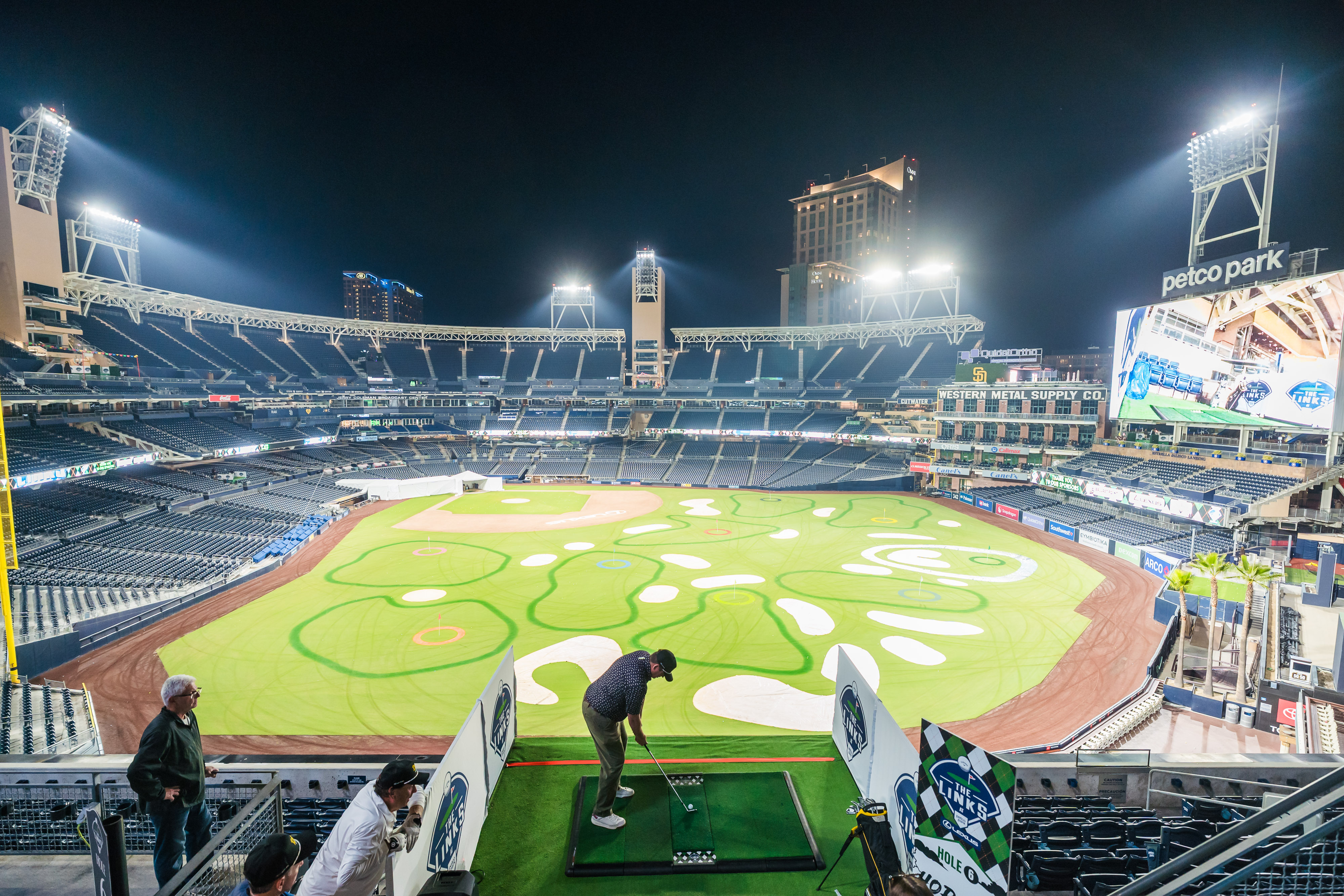 Indoor golf setup inside an empty Petco Park baseball stadium at night, people playing golf.