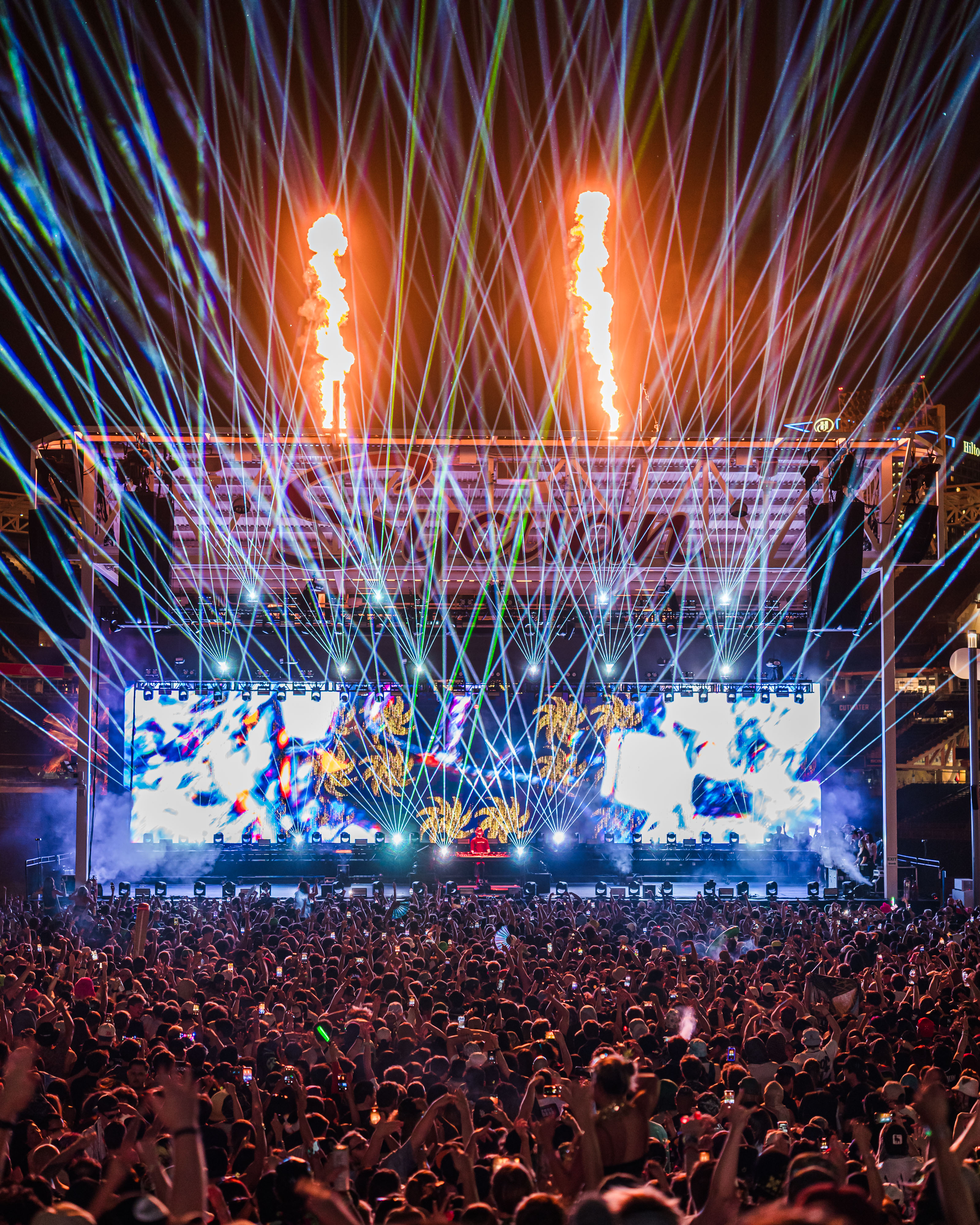 Bright laser lights and flames erupting above a stadium structure at night.
