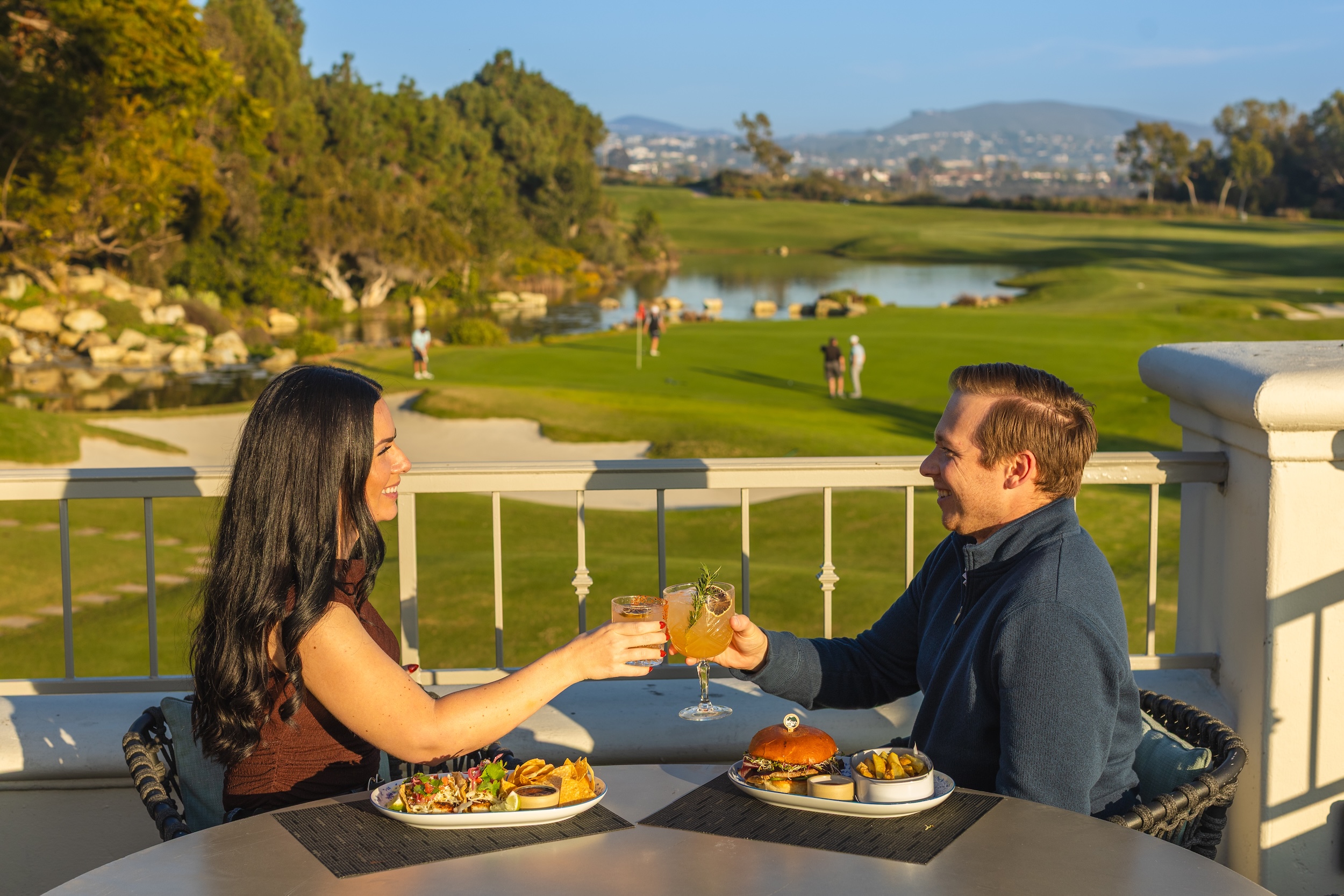 Couple clinking glasses over lunch at an outdoor table overlooking a golf course on a sunny day.