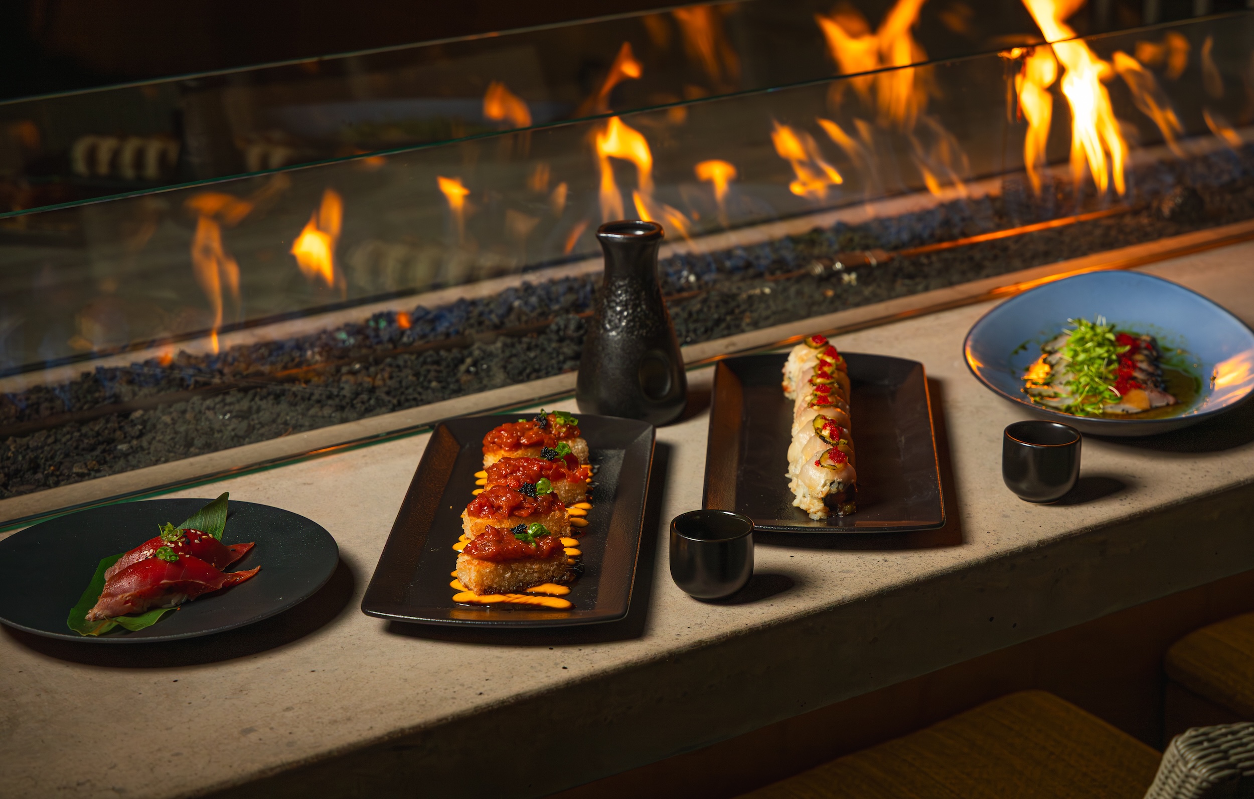 Assorted sushi dishes and sake set on a table by a modern indoor fire feature.