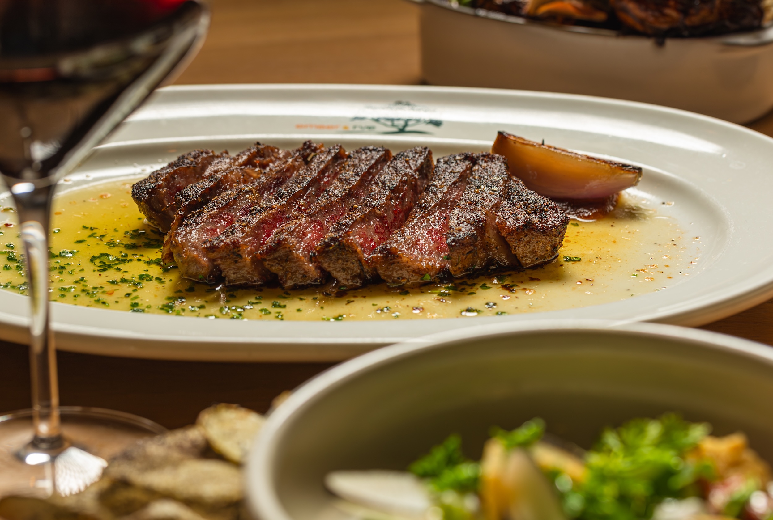 Sliced medium-rare steak on white plate with sauce, accompanied by salad and red wine glass.