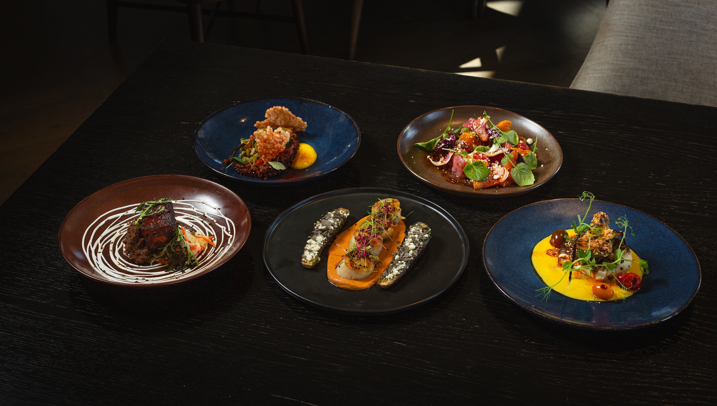 Five gourmet plated dishes arranged on a dark wooden table in a dimly lit setting.
