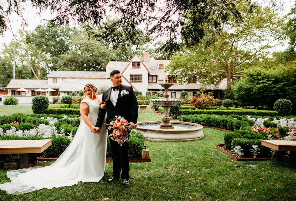Bride and groom standing together in a garden with a fountain, in front of a large house.