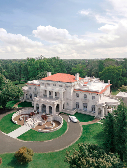 Large, white mansion with red roof, circular driveway, surrounded by trees under a partly cloudy sky.