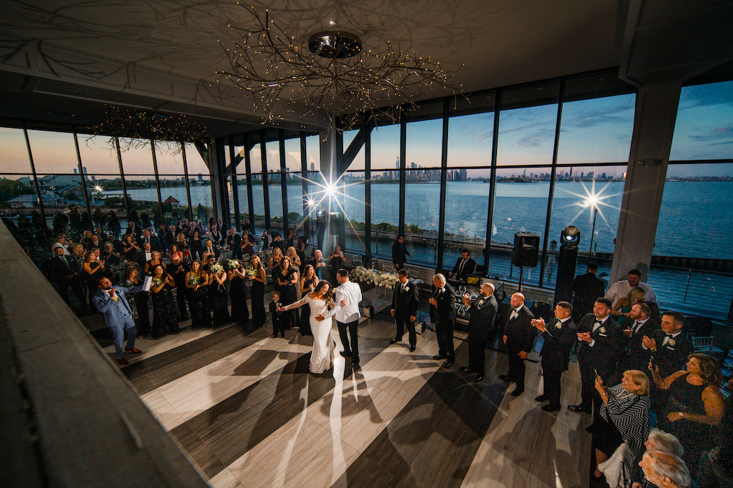 Wedding couple dancing surrounded by guests in a waterfront venue with large windows at sunset.