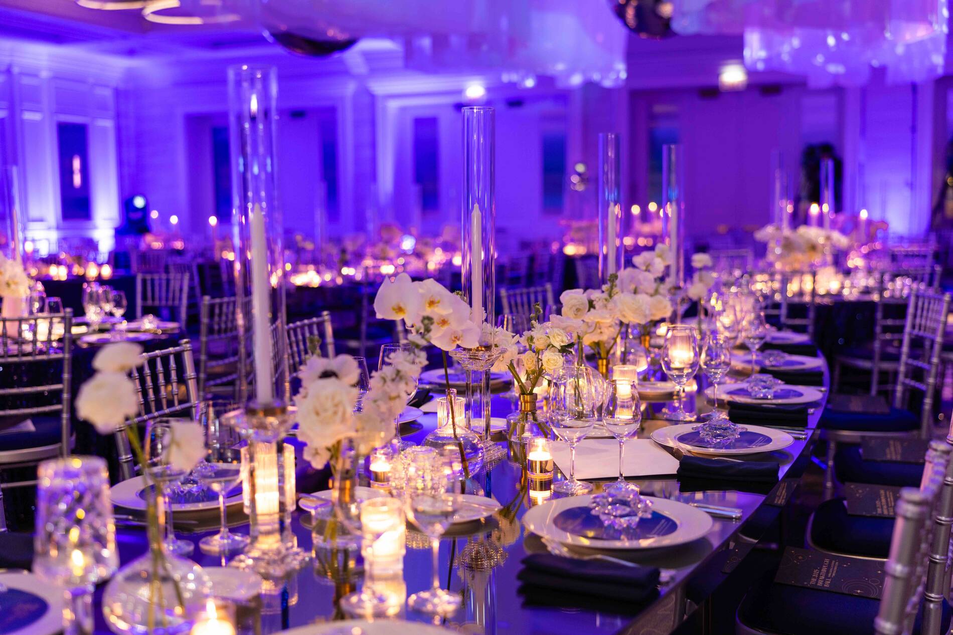 Elegant banquet hall with long tables, floral centerpieces, and soft purple lighting.