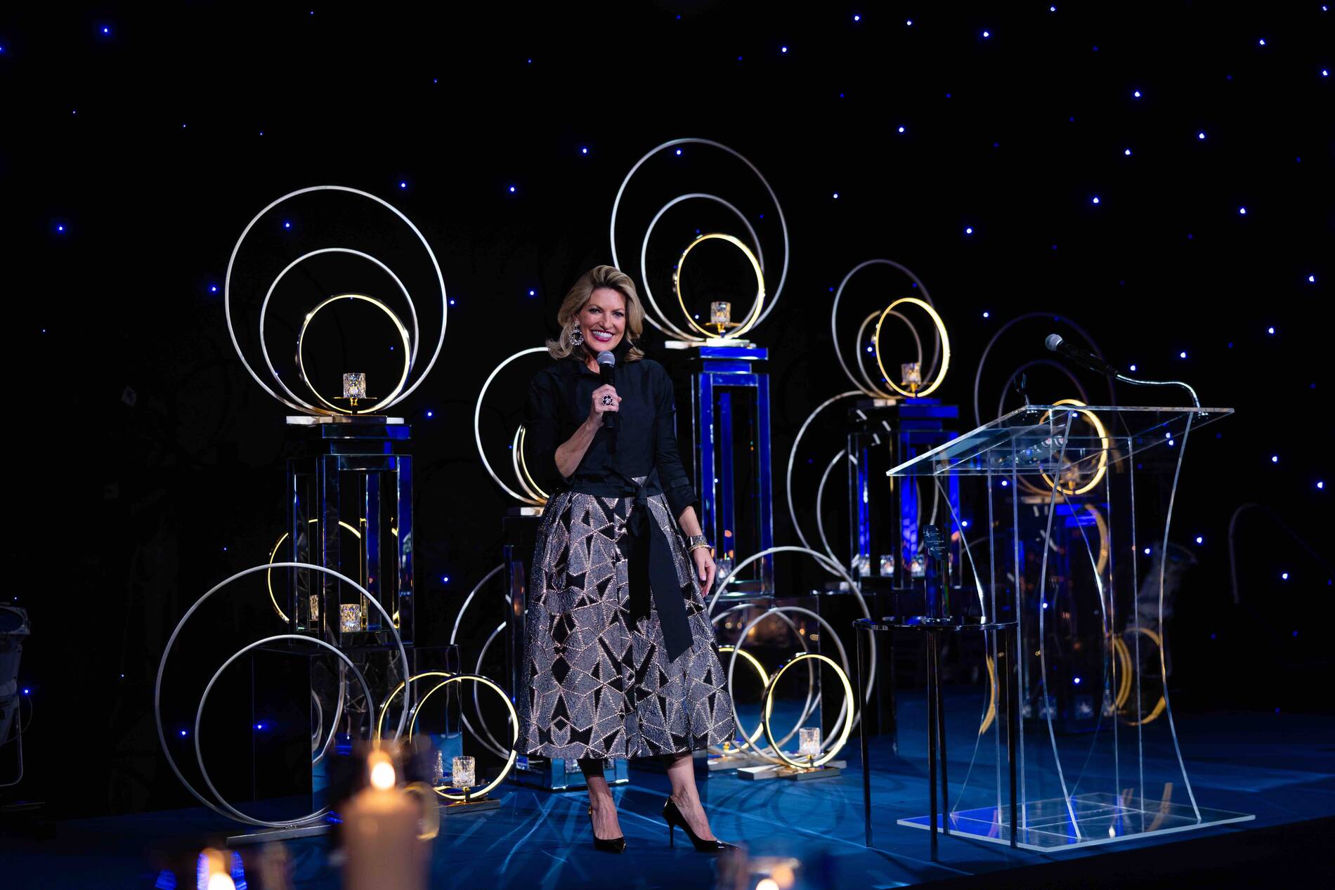 Woman speaking on stage with decorative circular lights and a starry background at an event.