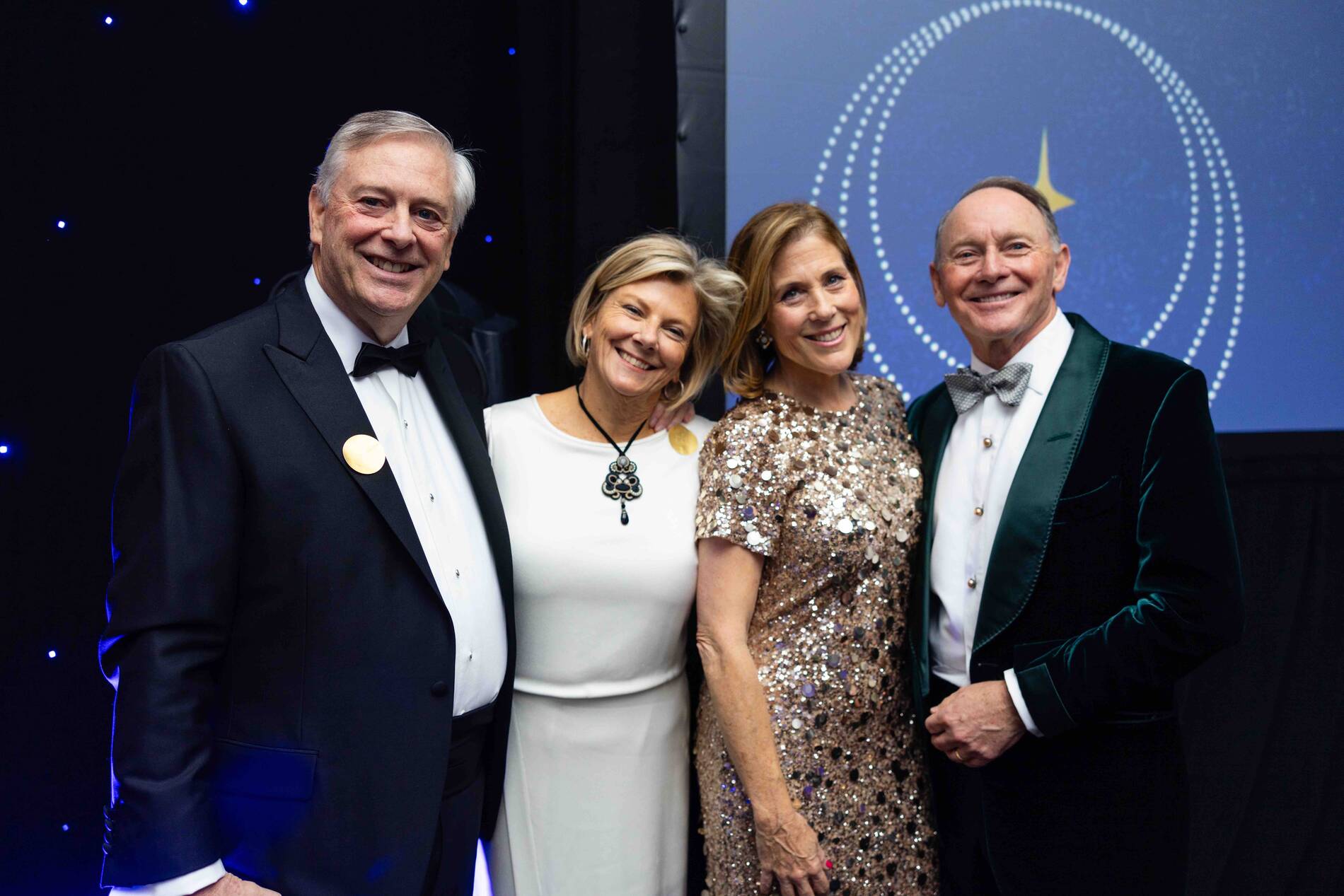 Four adults dressed formally, smiling and posing at an event with a decorated backdrop.