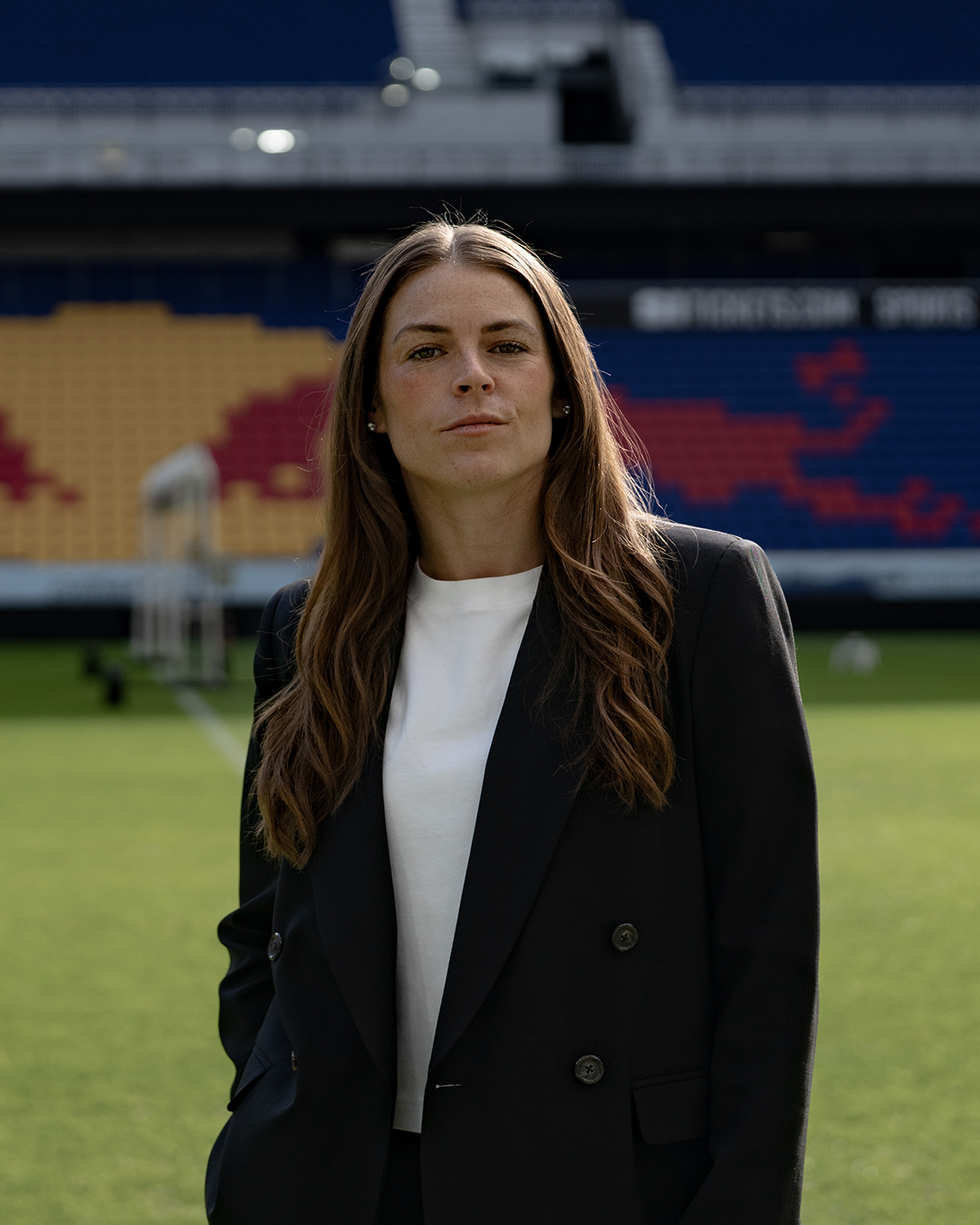 Portrait of Kelley O’Hara standing on a soccer field in a black blazer, with stadium seating blurred in the background.