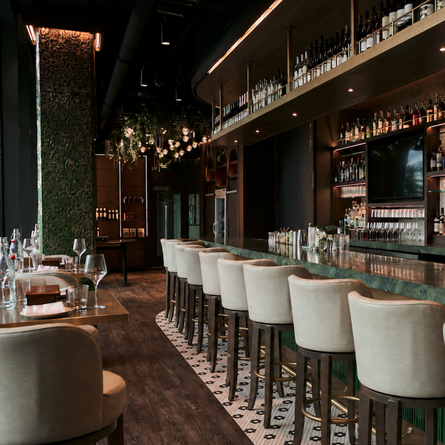 Elegant empty bar with beige stools, wooden tables, decorative green wall, and overhead wine bottle display.