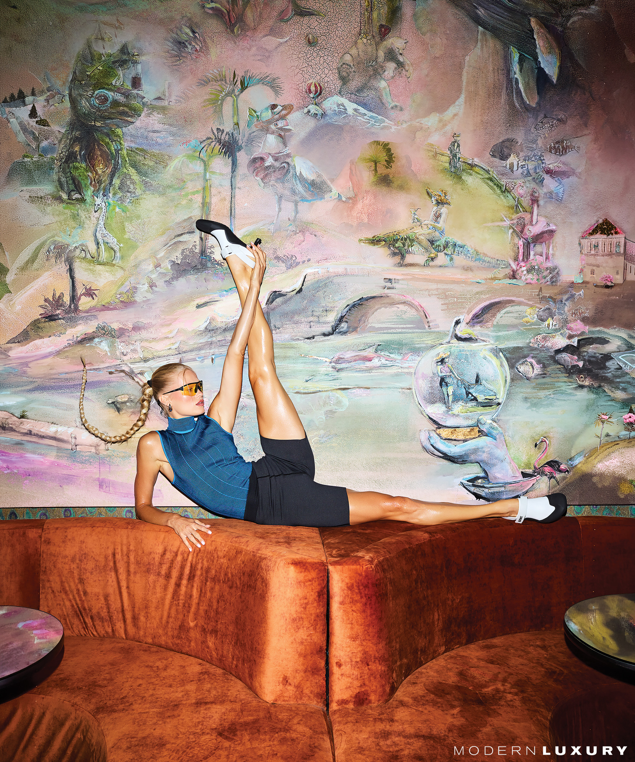 Vita Sidorkina-Morabito with long braided hair poses on rust-colored couch with colorful surreal mural background wearing an Hermès top and shorts.