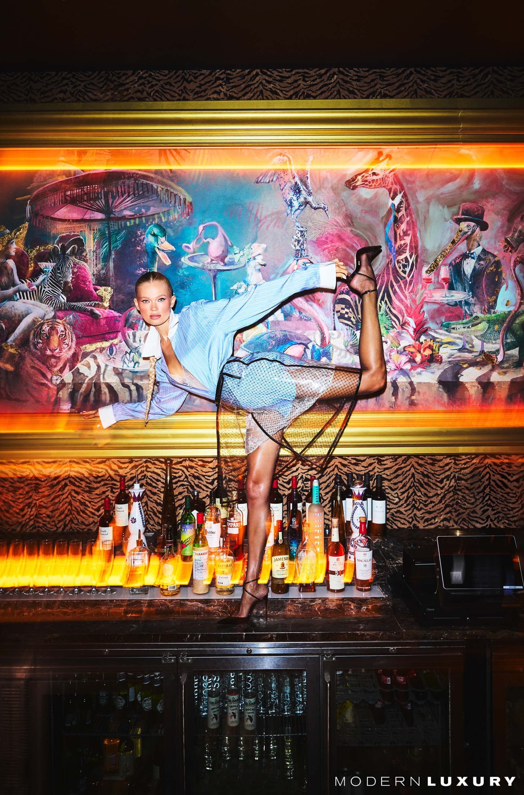 Vita Sidorkina-Morabito poses on a bar at Mary Lou's Miami wearing a Tom Ford blue outfit. She is holding her leg and leaning foward.