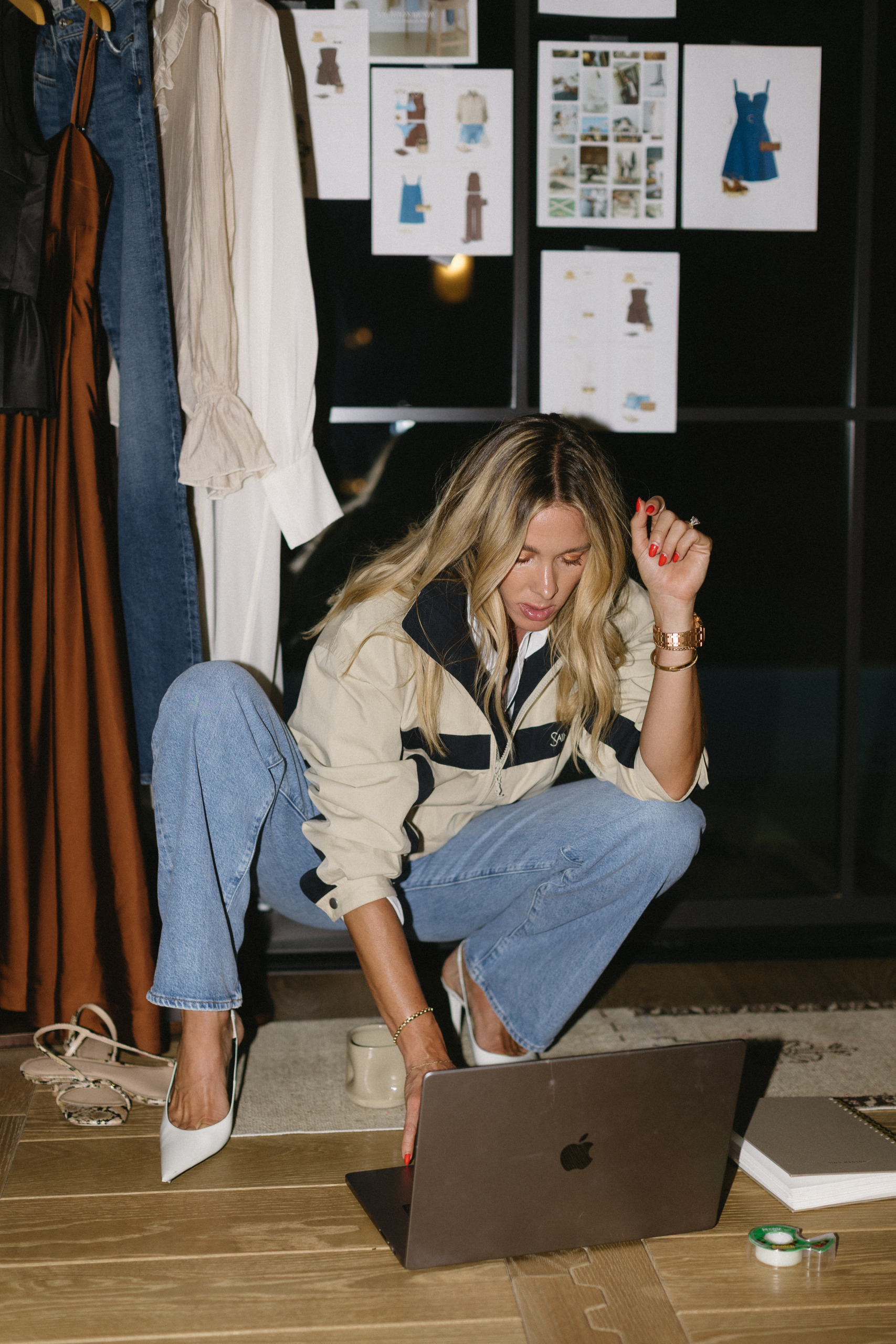 Woman with blonde hair wearing a jacket and jeans, working on a laptop in a room with clothes and fashion sketches.