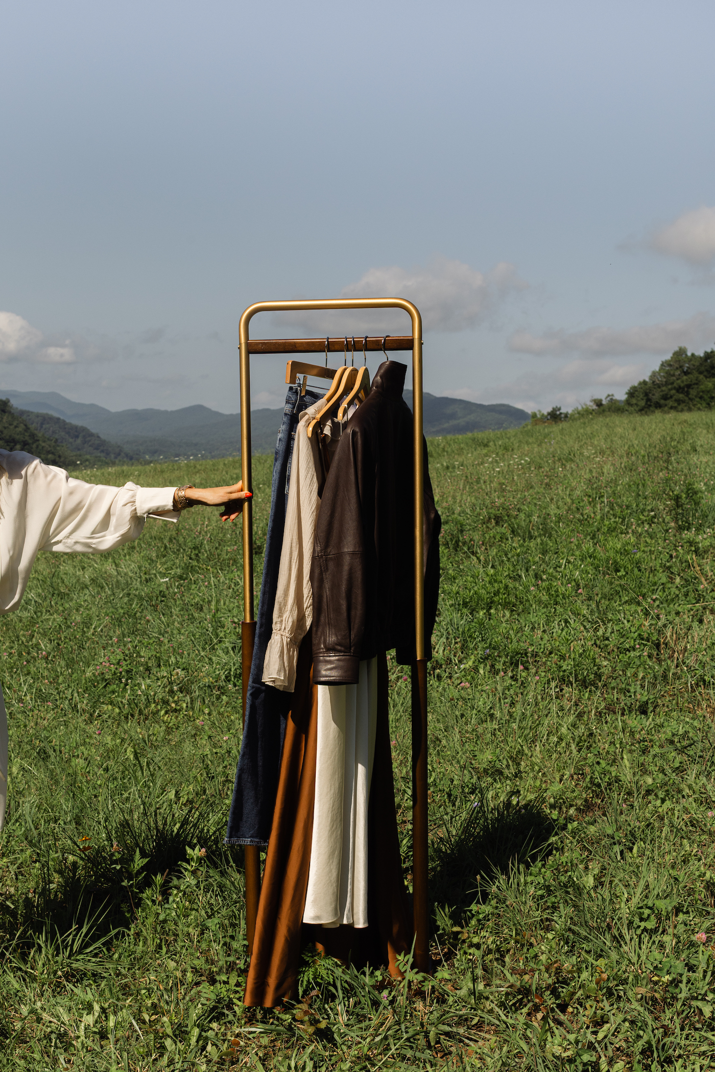 Clothing rack with jackets and pants in a grassy field, hand holding the rack on the left.